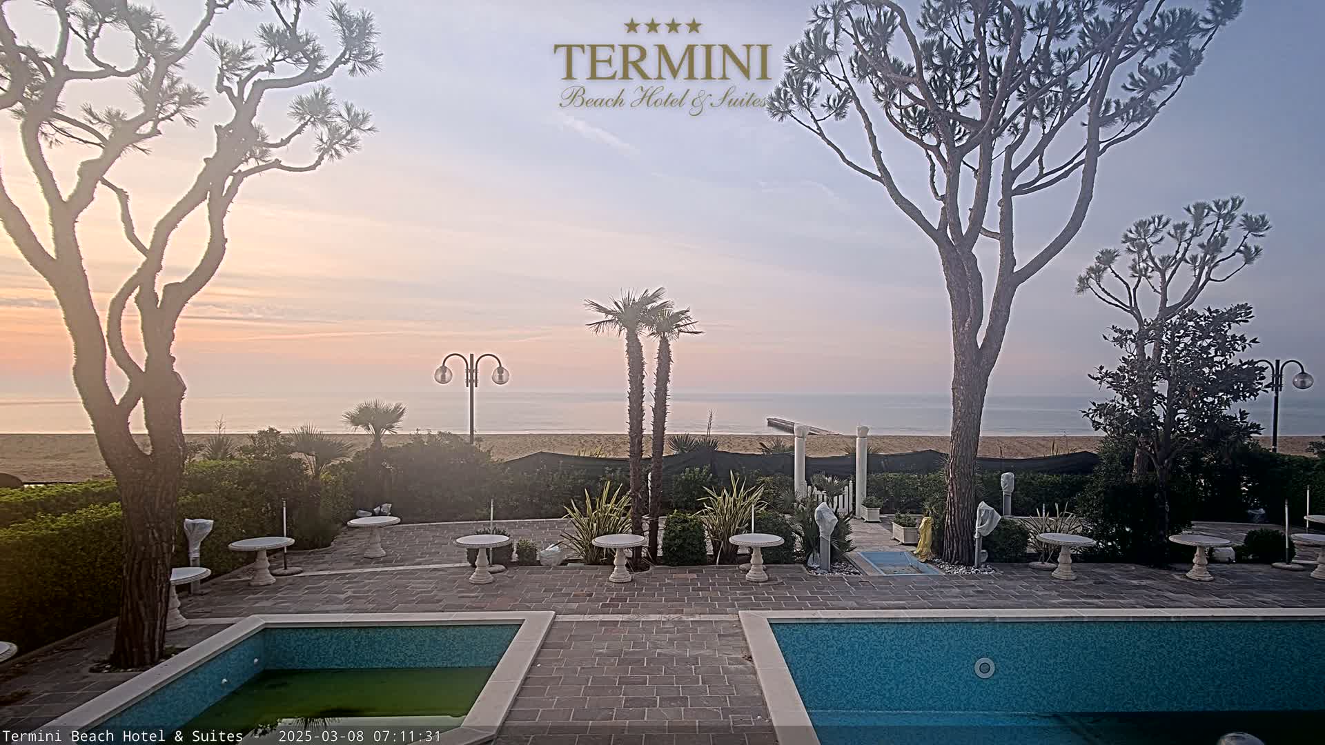 A calm ocean is visible in the distance beyond a patio area with two swimming pools, several small tables, and palm trees under a pale sunrise.