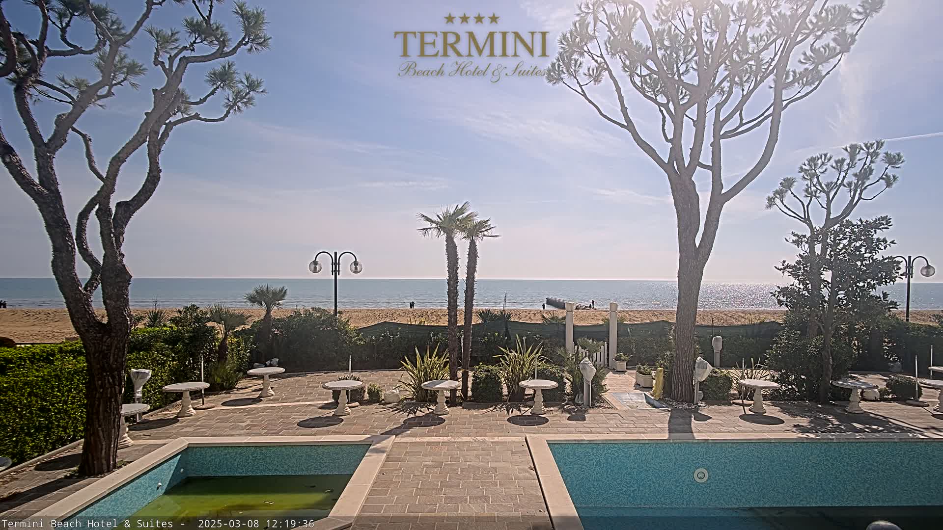 A sunny outdoor scene shows a patio area with two swimming pools, some small tables, and landscaping, all overlooking a sandy beach and calm ocean.