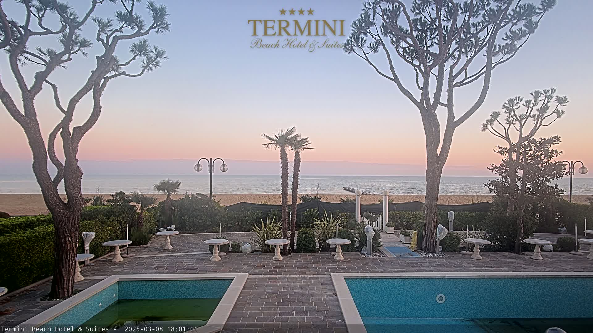 A tranquil outdoor scene at sunset shows a courtyard with two swimming pools, several small tables, and lush landscaping overlooking a calm ocean beach.