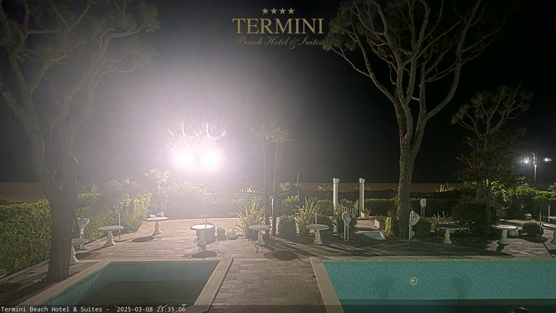A nighttime view of a patio area with two swimming pools, several small tables, and trees, illuminated by bright lights in hazy conditions.