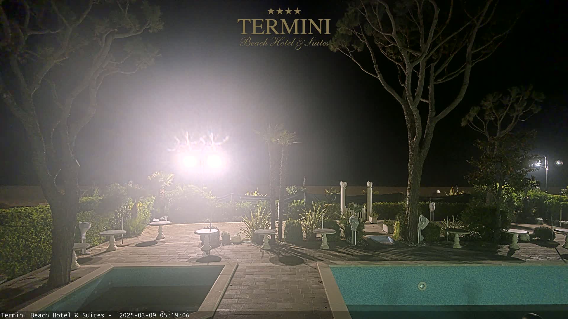 A nighttime scene shows a swimming pool surrounded by trees, small tables, and landscaping, illuminated by bright lights.