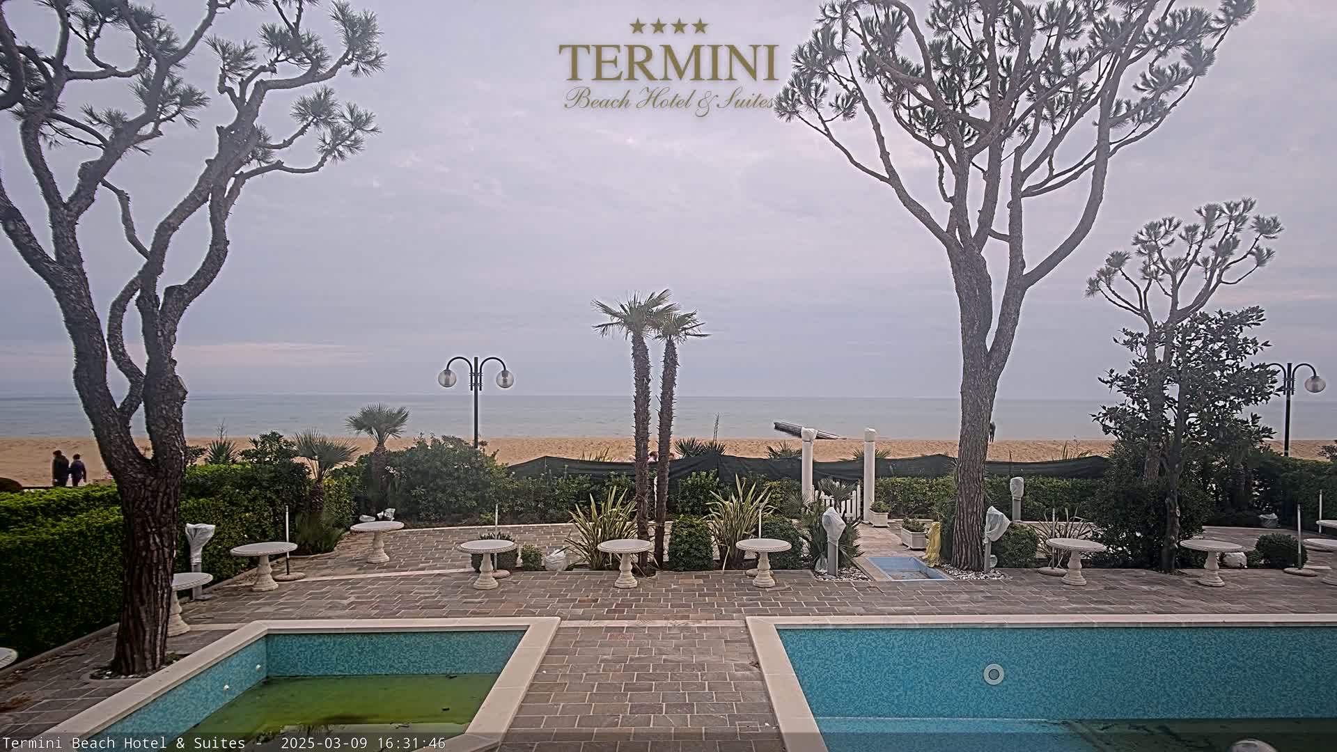 A patio with two small pools overlooks a calm ocean and sandy beach under an overcast sky.