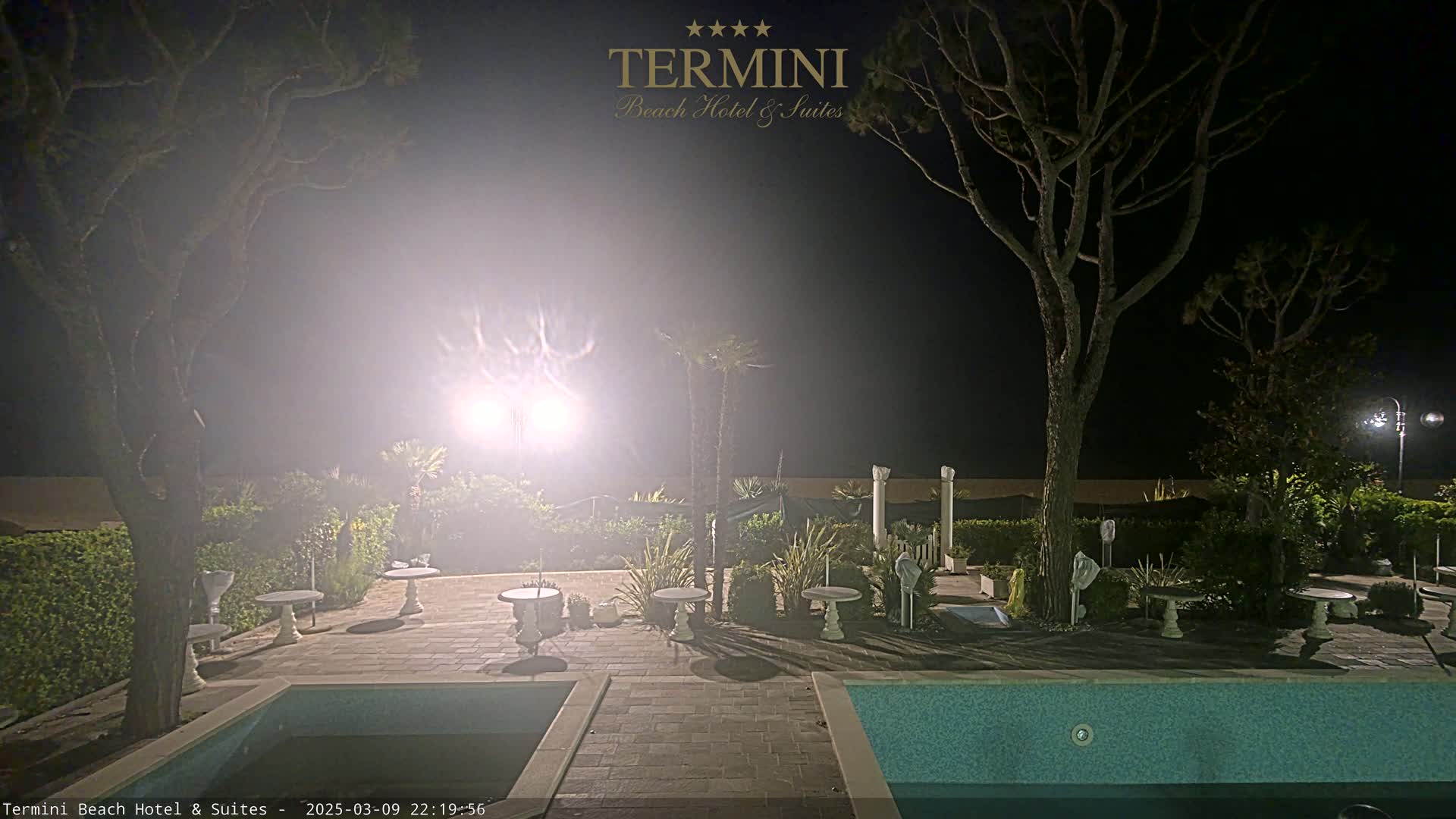 A hazy night scene shows an outdoor patio area with a swimming pool, several small tables, and tall trees surrounding the area.