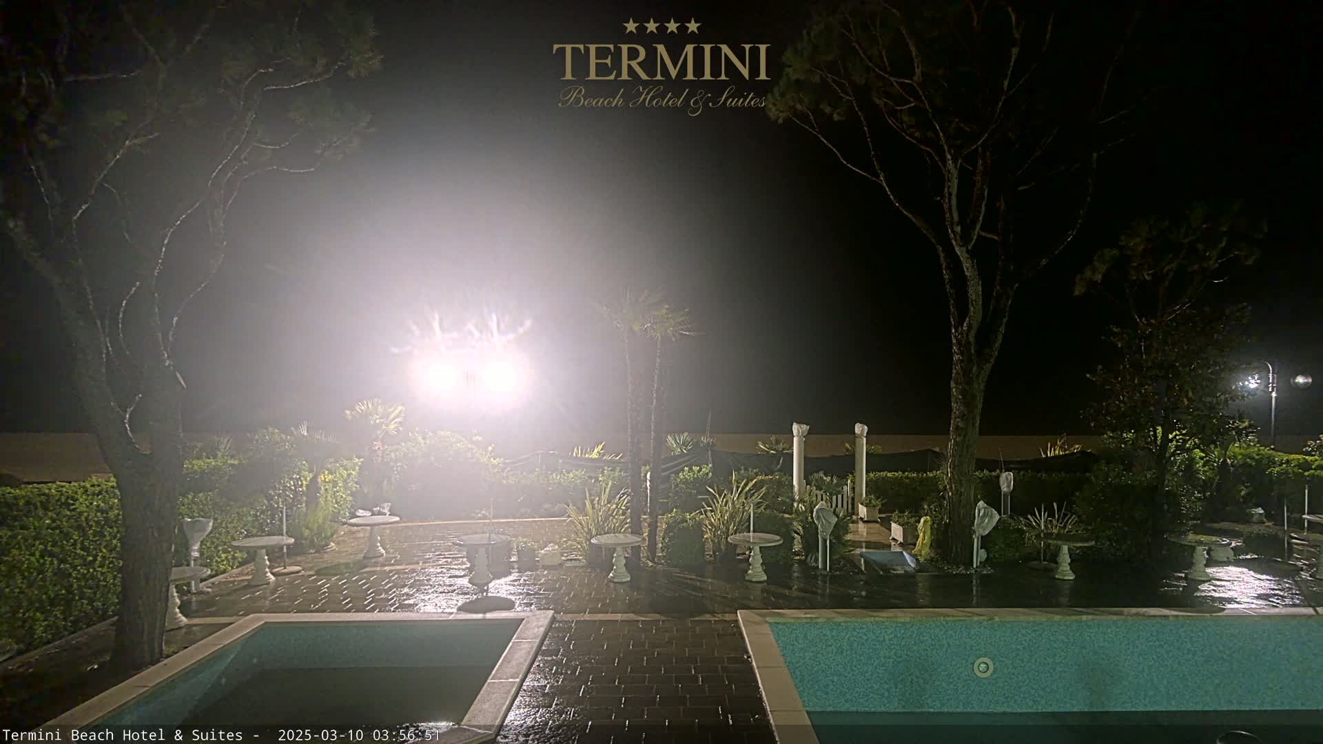 A nighttime view of a patio area with two swimming pools, surrounded by trees and landscaping, under hazy conditions.