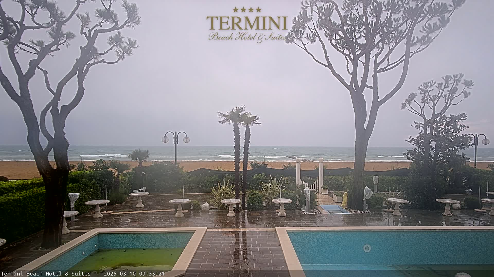 A wet patio area with two pools overlooks a beach and ocean under an overcast sky.