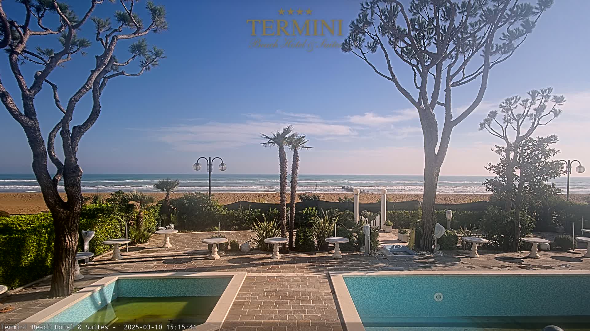 A patio with two swimming pools overlooks a sandy beach and calm ocean under a clear blue sky.
