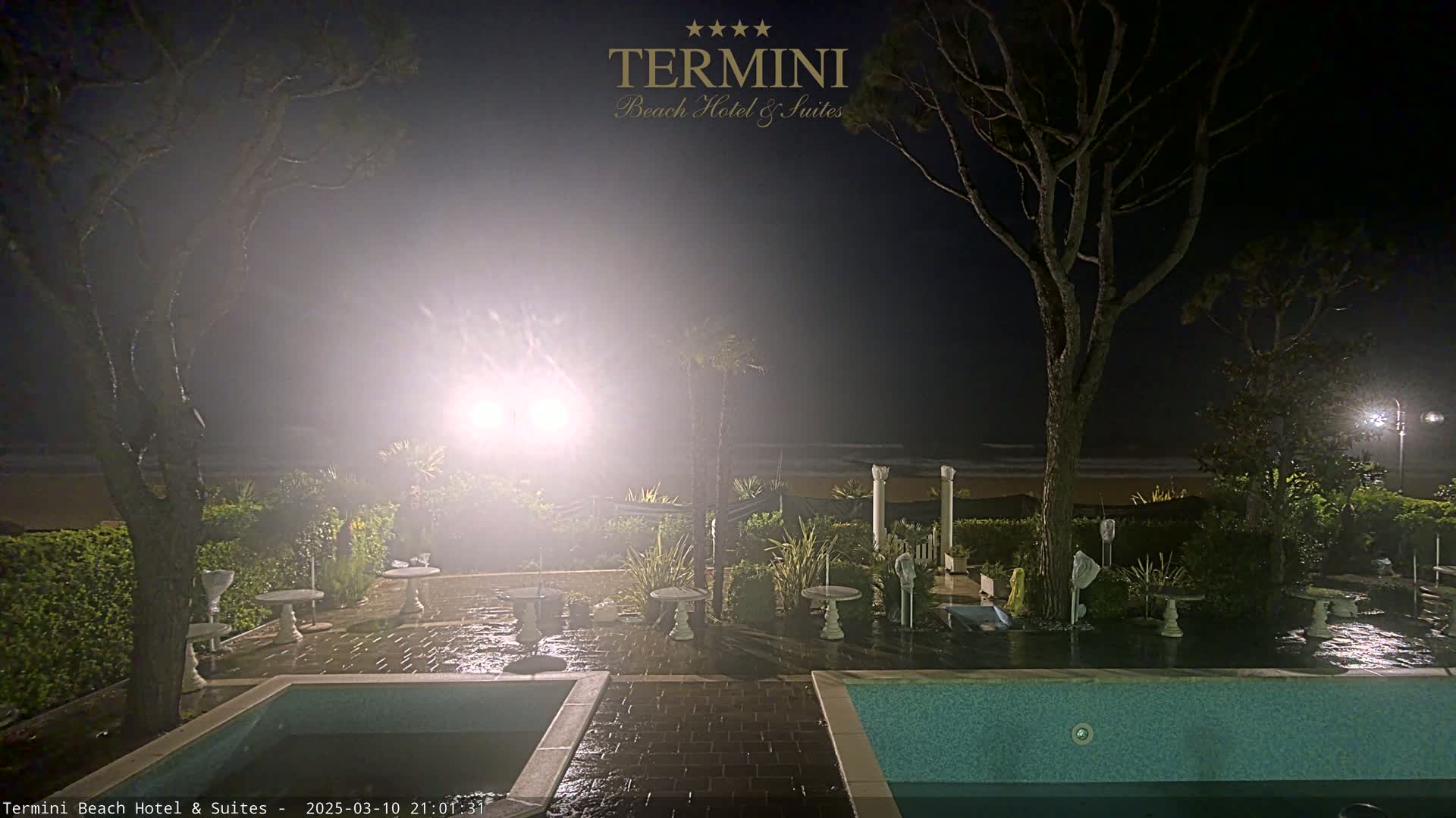 A nighttime view of a patio area with two swimming pools, outdoor furniture, and trees, overlooking a beach under hazy conditions.
