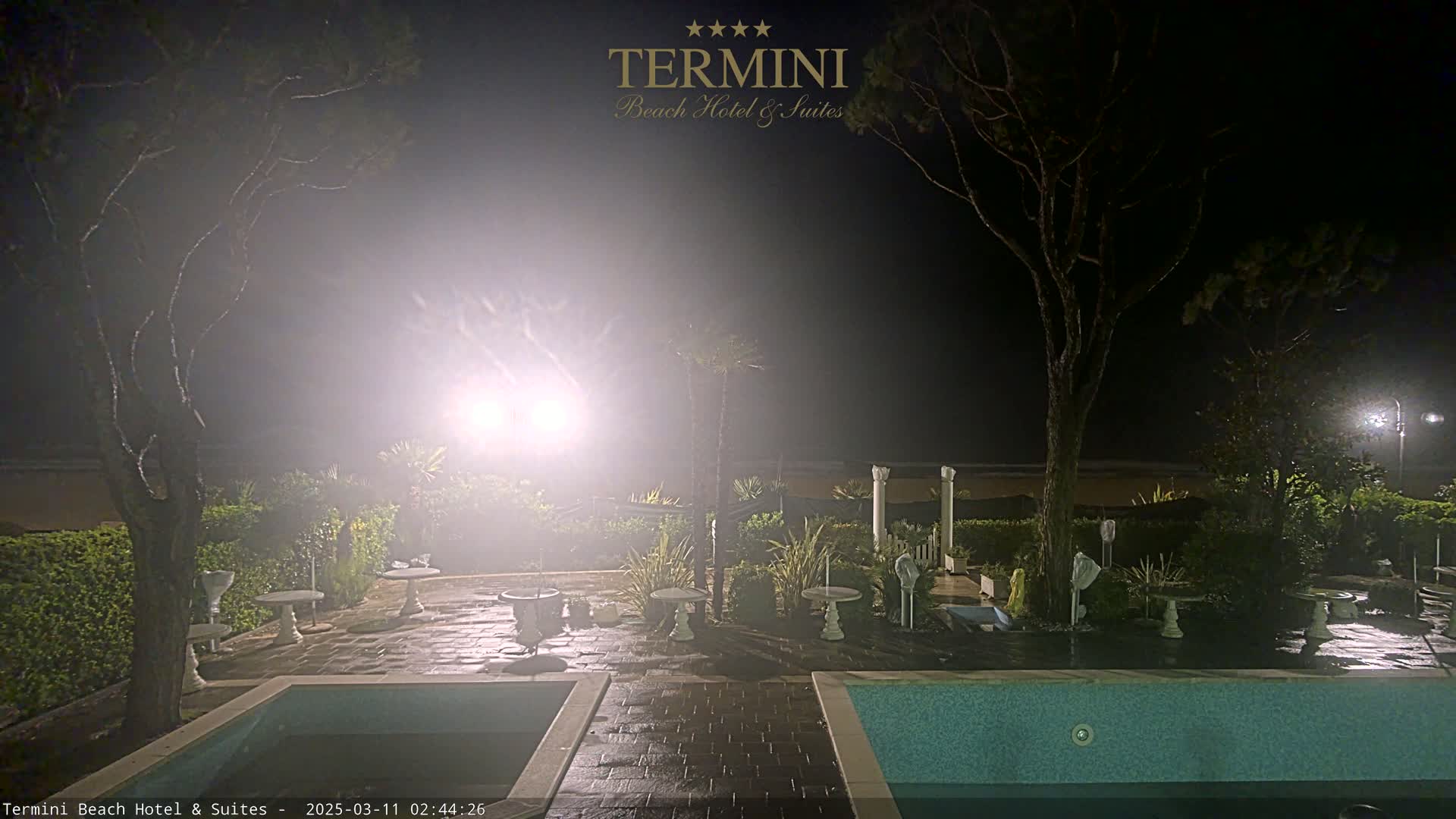 A nighttime view of a hotel patio area with two swimming pools, several small tables and urns, and trees, illuminated by bright lights in foggy or hazy conditions.