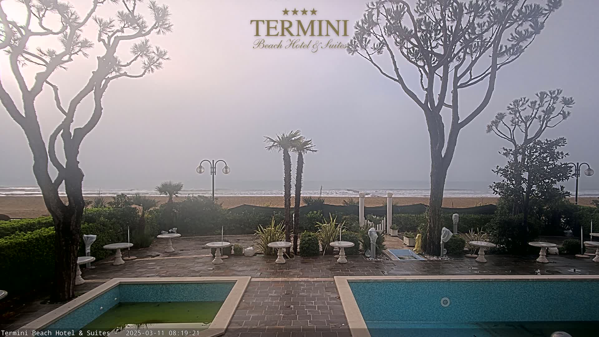 A hazy, overcast day reveals a sandy beach and ocean viewed from a hotel courtyard with two swimming pools, several small tables, and palm and pine trees.