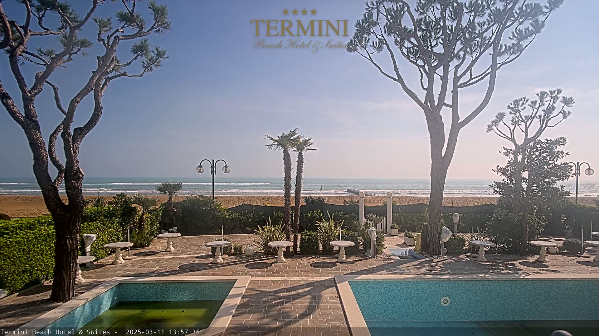 A patio with two pools overlooks a sandy beach and calm ocean under a clear, sunny sky.