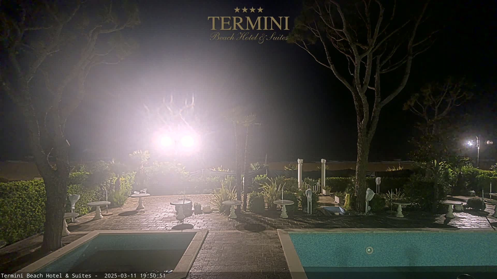 A nighttime scene shows a hazy outdoor patio area with two pools, several small tables and chairs, and trees surrounding it.