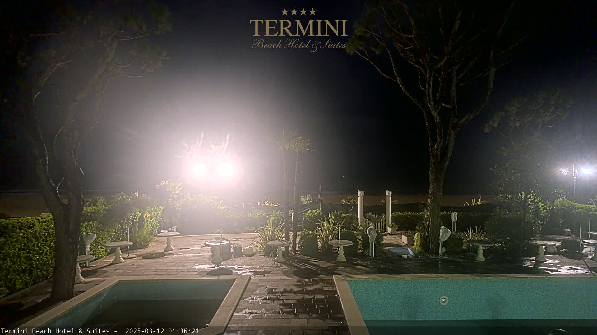 A nighttime view of a patio area with a swimming pool, surrounded by trees and small tables, under bright lights, near a beach.