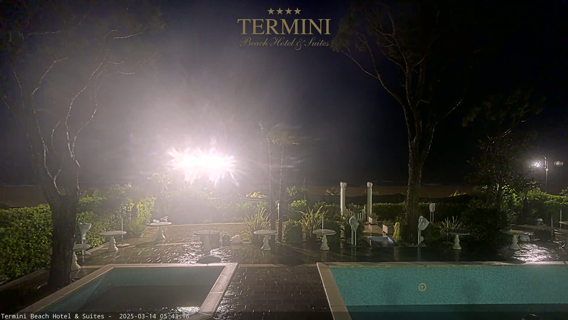A nighttime view of a resort's outdoor patio area, featuring two pools, landscaping, and some outdoor furniture, with hazy or foggy conditions and lights illuminating the scene.