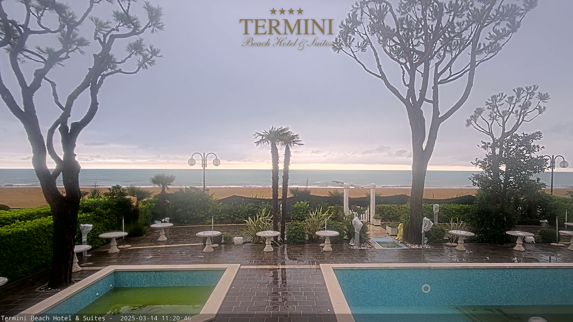 An overcast sky overlooks a sandy beach and calm ocean from a patio with two pools, tables, and landscaping, including palm trees and pine trees.