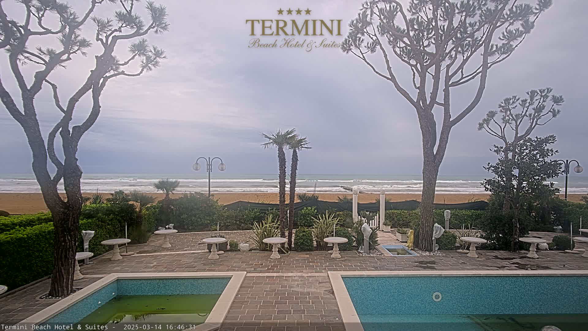 An overcast day shows a sandy beach with a calm ocean viewed from a hotel patio with two swimming pools, landscaping, and several trees.