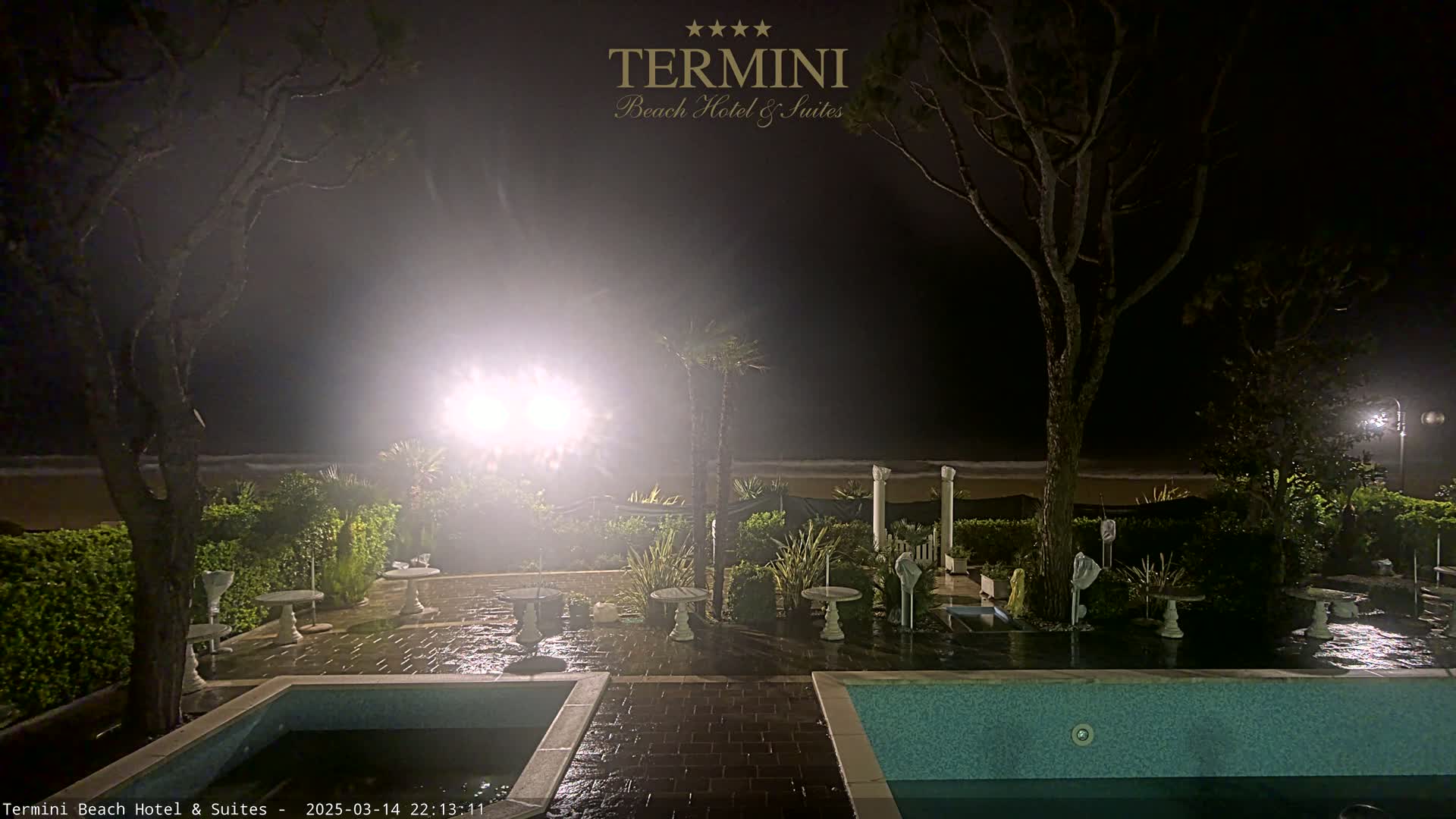 A nighttime view of a resort pool area with wet patio furniture near a beach under hazy conditions.