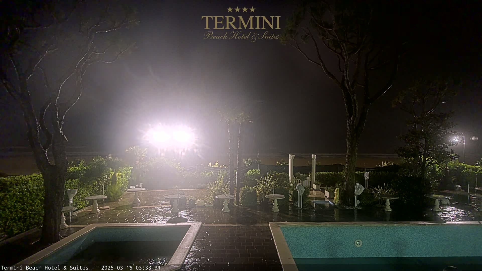 A nighttime view of a patio area with two pools, several small tables and trees, all under a hazy, misty atmosphere near a dimly lit beach.