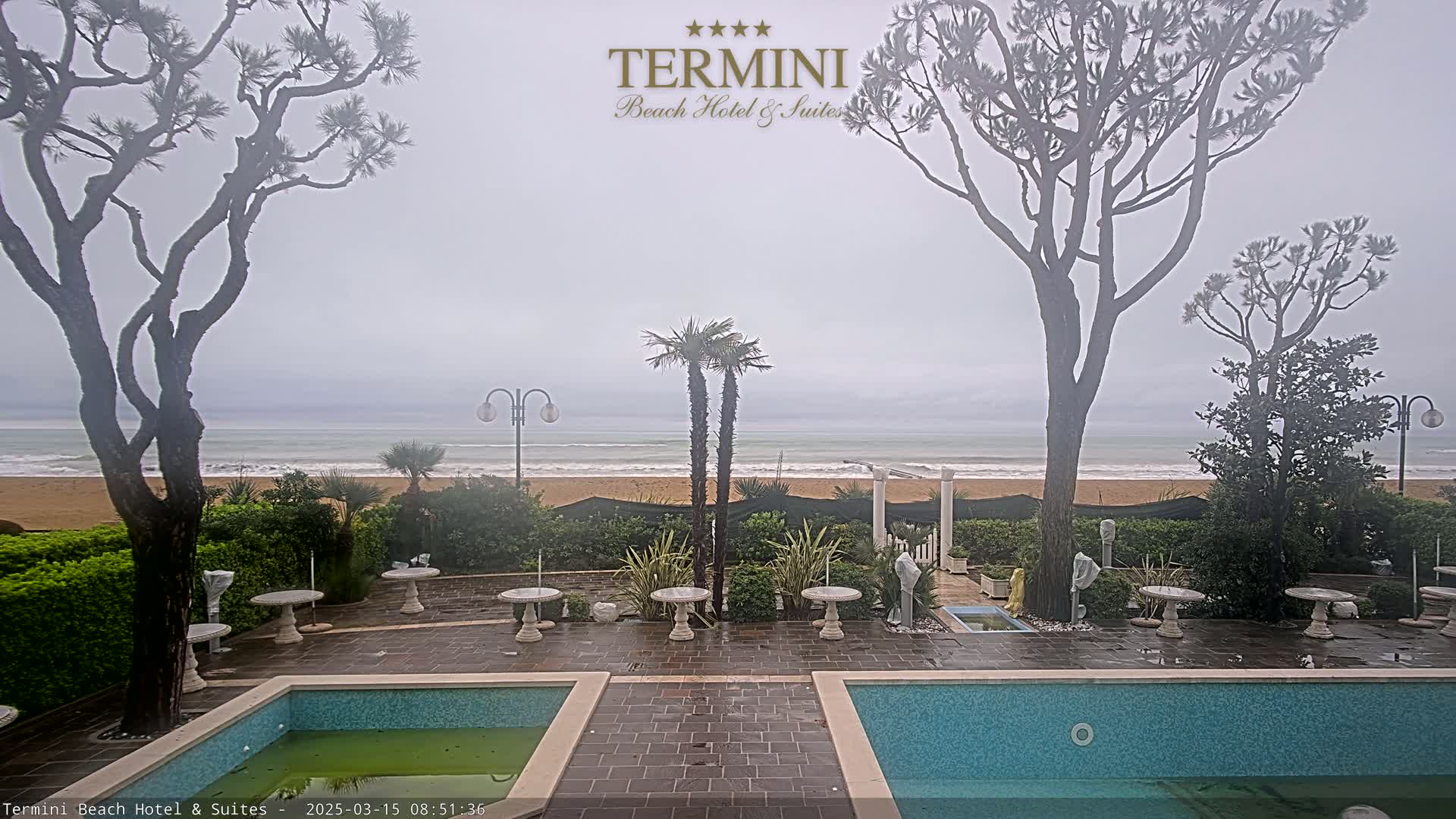 An overcast day shows a patio area with two pools overlooking a beach and ocean with small waves.