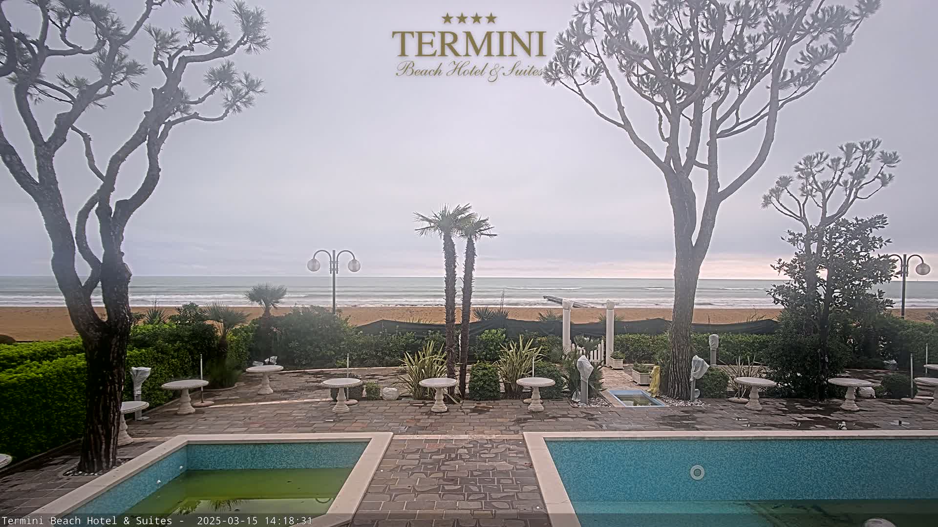 A patio with two pools overlooks a beach and ocean under an overcast sky.