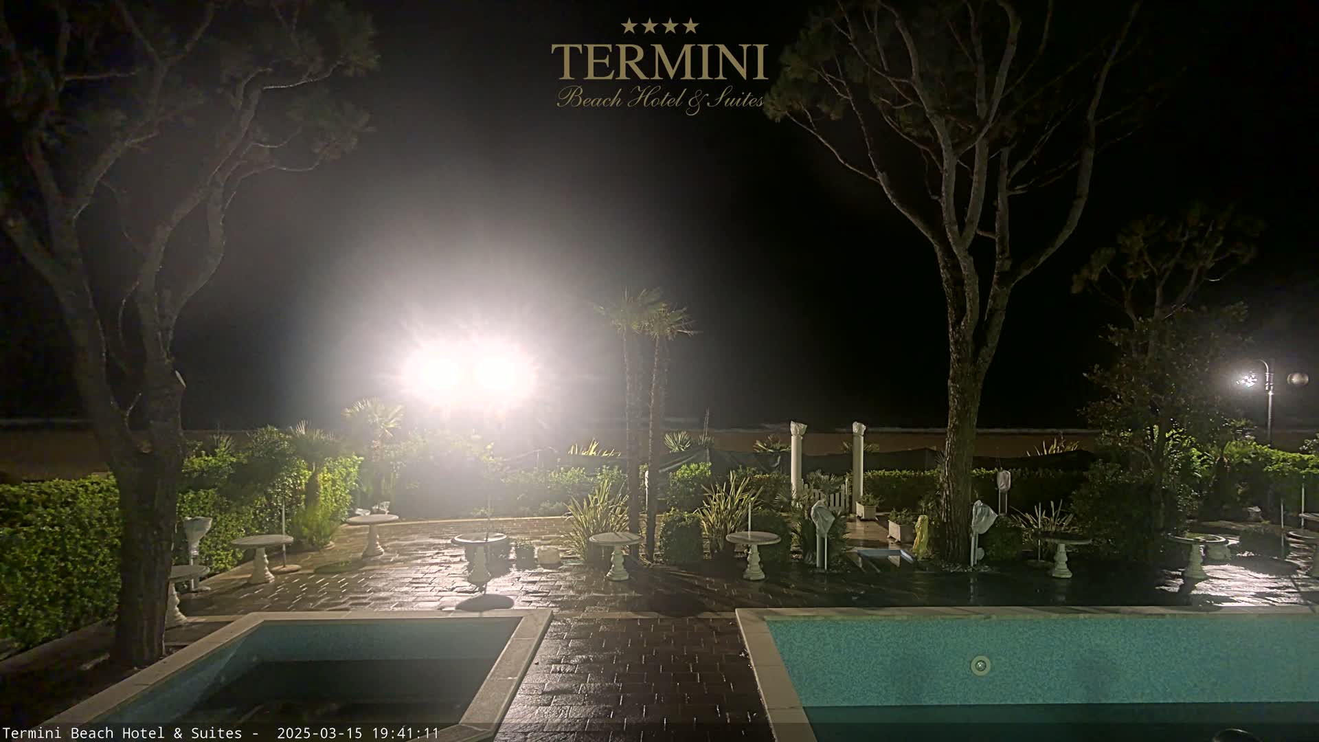 A nighttime view of a patio area with a pool, featuring small tables and potted plants, illuminated by artificial light.