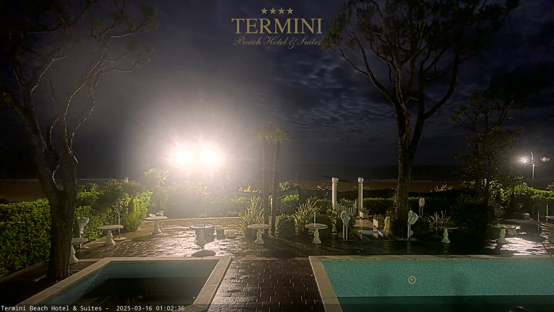 An outdoor patio area at night with a pool, landscaping, and some outdoor furniture is illuminated by lights, overlooking a dark ocean under a partly cloudy sky.