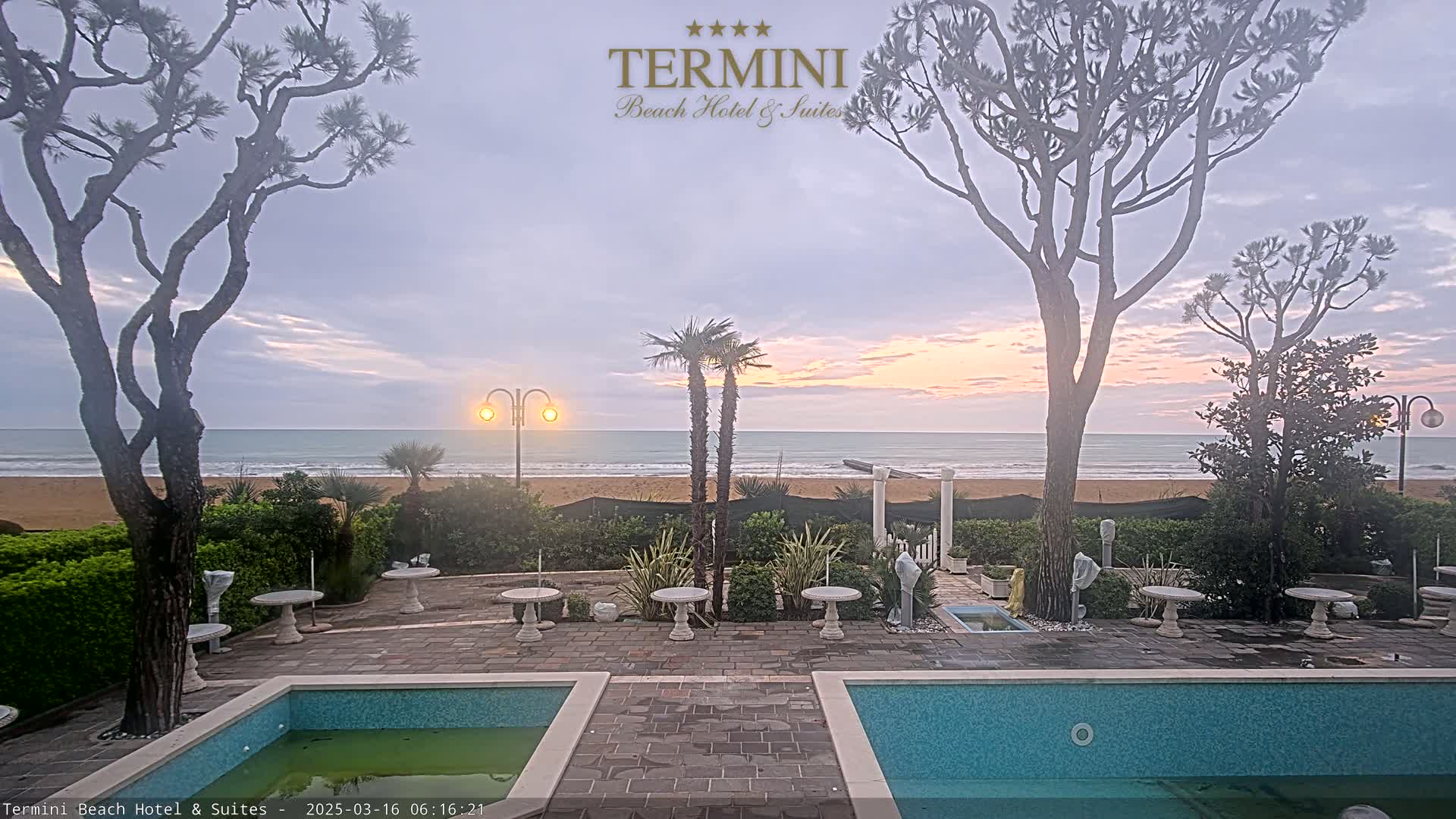 A calm ocean with a sandy beach is visible from a patio area with two swimming pools, several tables, and trees, during a sunrise or sunset with overcast skies.