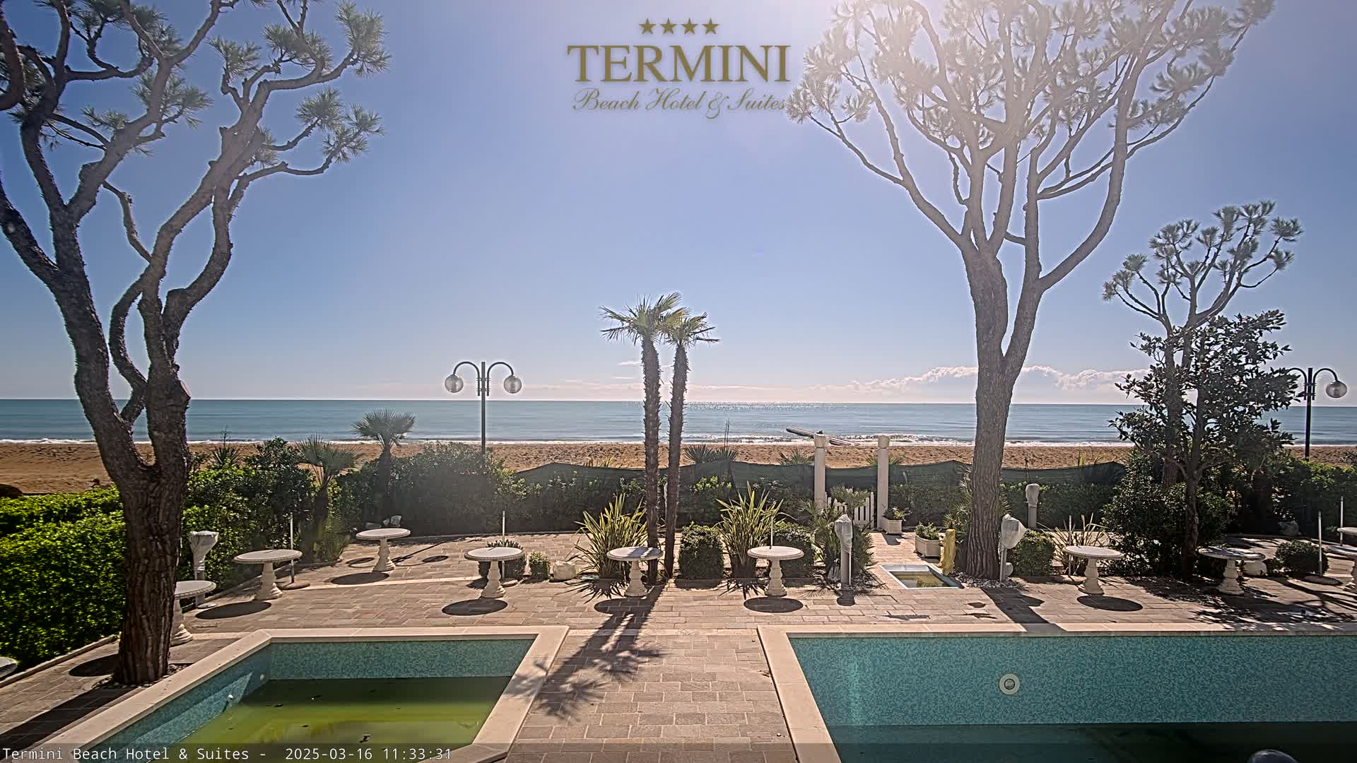 A sunny outdoor patio scene with two swimming pools, several small tables and chairs, and palm trees, overlooking a calm ocean beach.