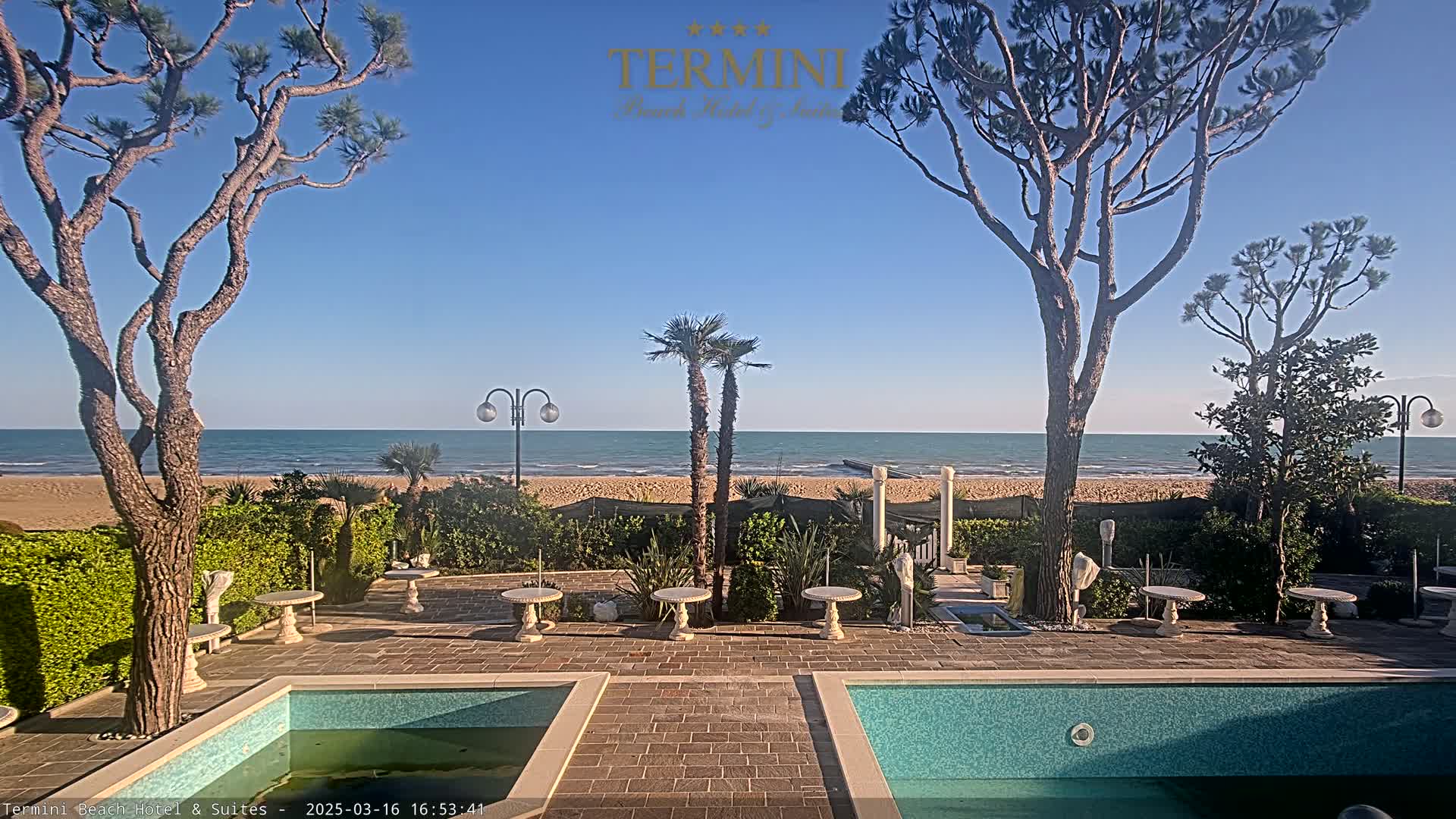 A patio with two swimming pools overlooks a calm ocean beach on a sunny day.