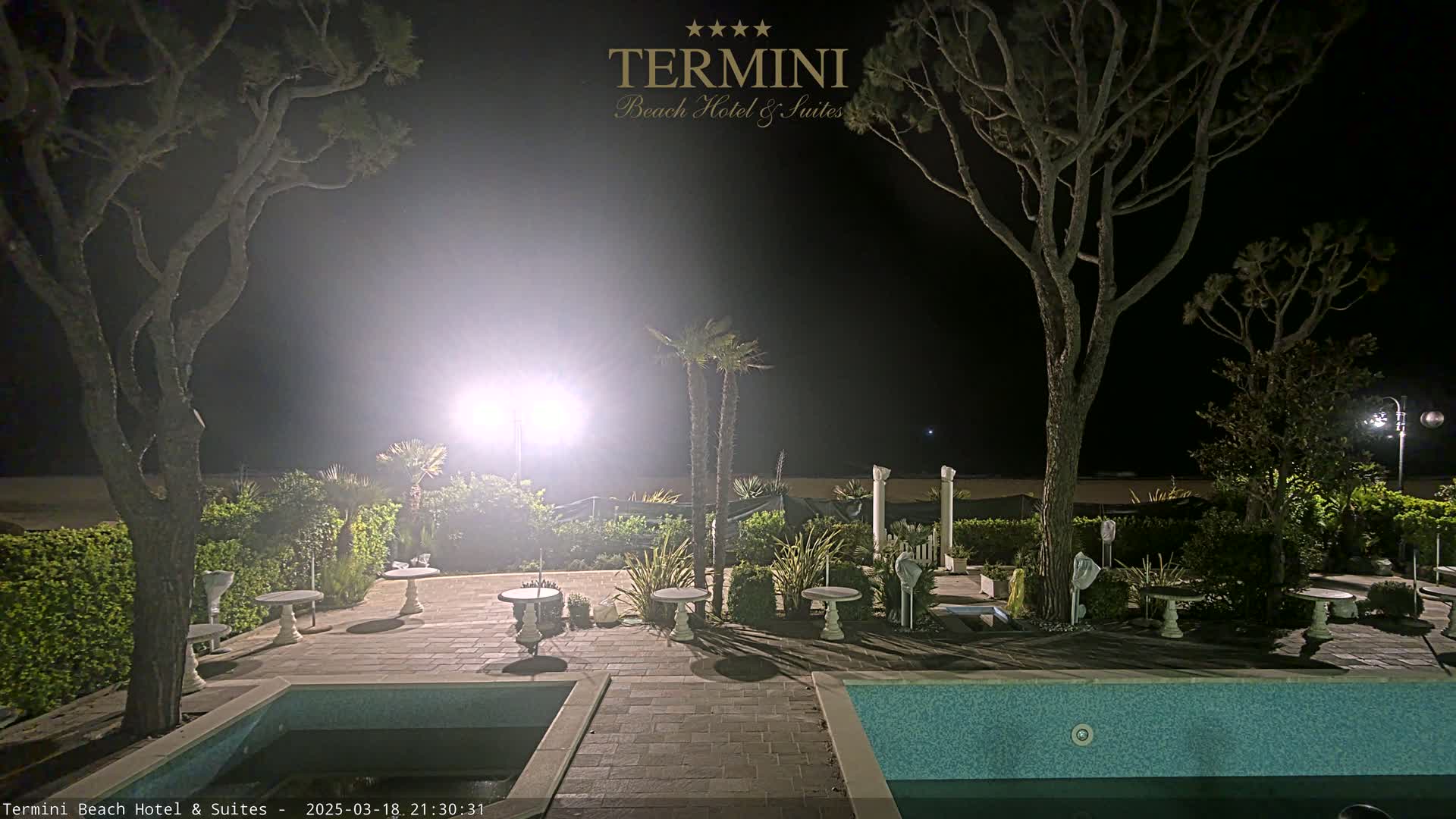 A nighttime scene shows a patio area with a swimming pool, several small tables, and trees surrounding a brightly lit space near a beach.