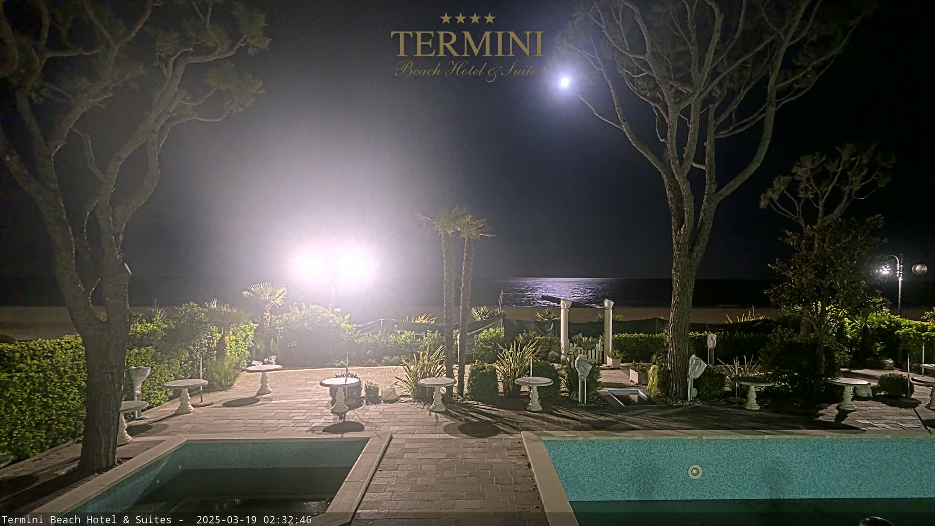 A moonlit night scene shows a patio area with two pools and small tables, surrounded by trees and hedges, overlooking a calm ocean.