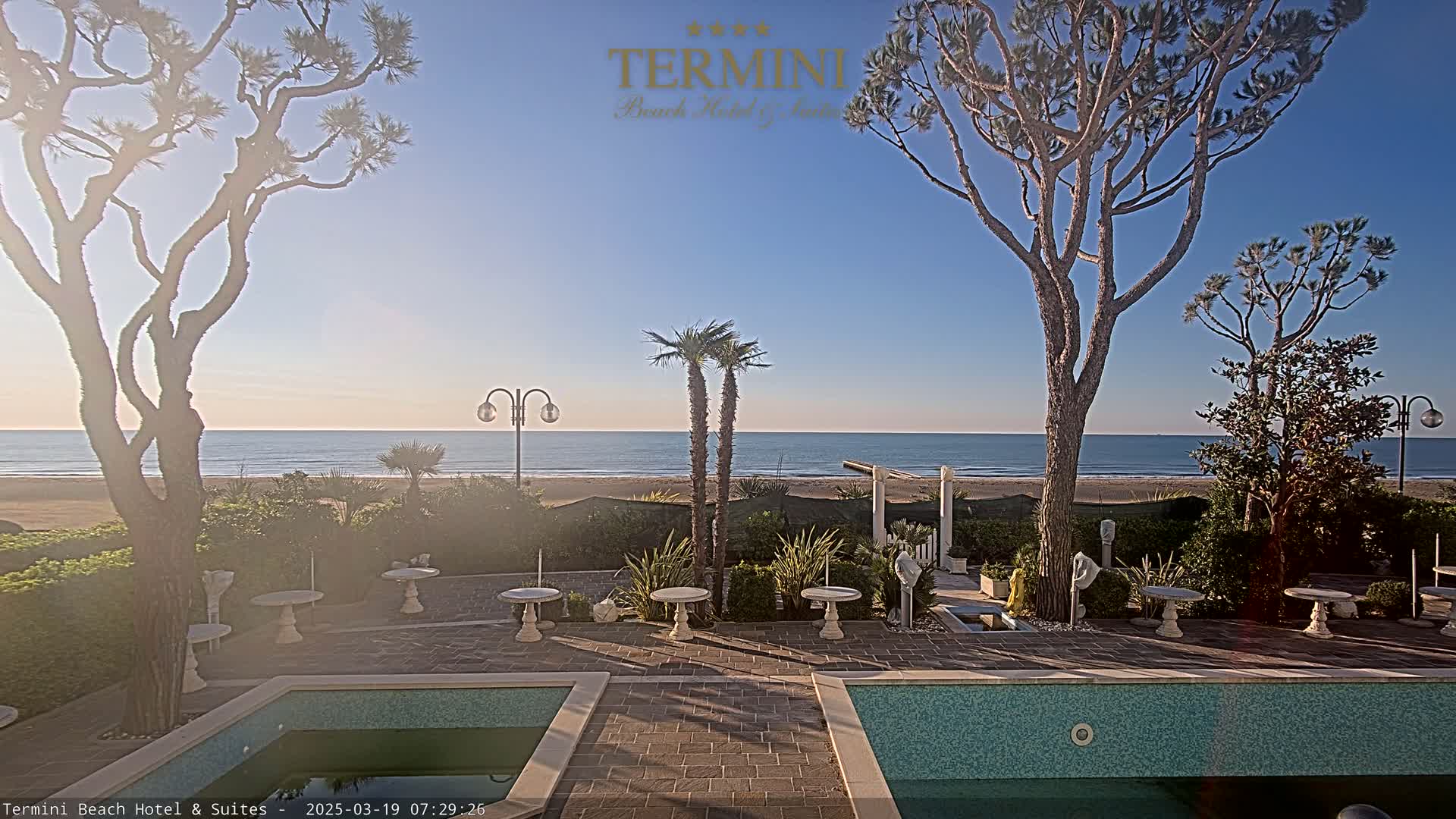 A view from a patio with two swimming pools overlooking a calm ocean under a clear, sunny sky.