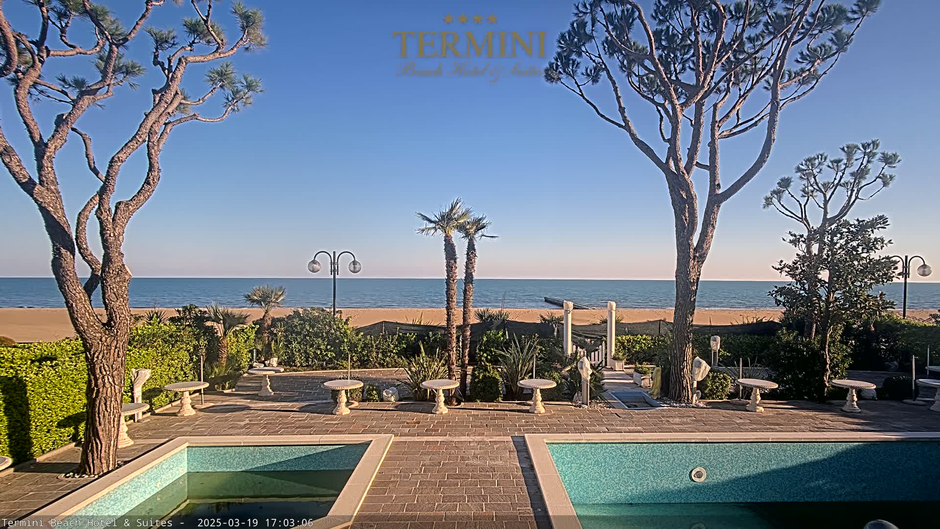 A calm ocean is visible beyond a patio area with two pools, several small tables, and tall trees under a clear blue sky.
