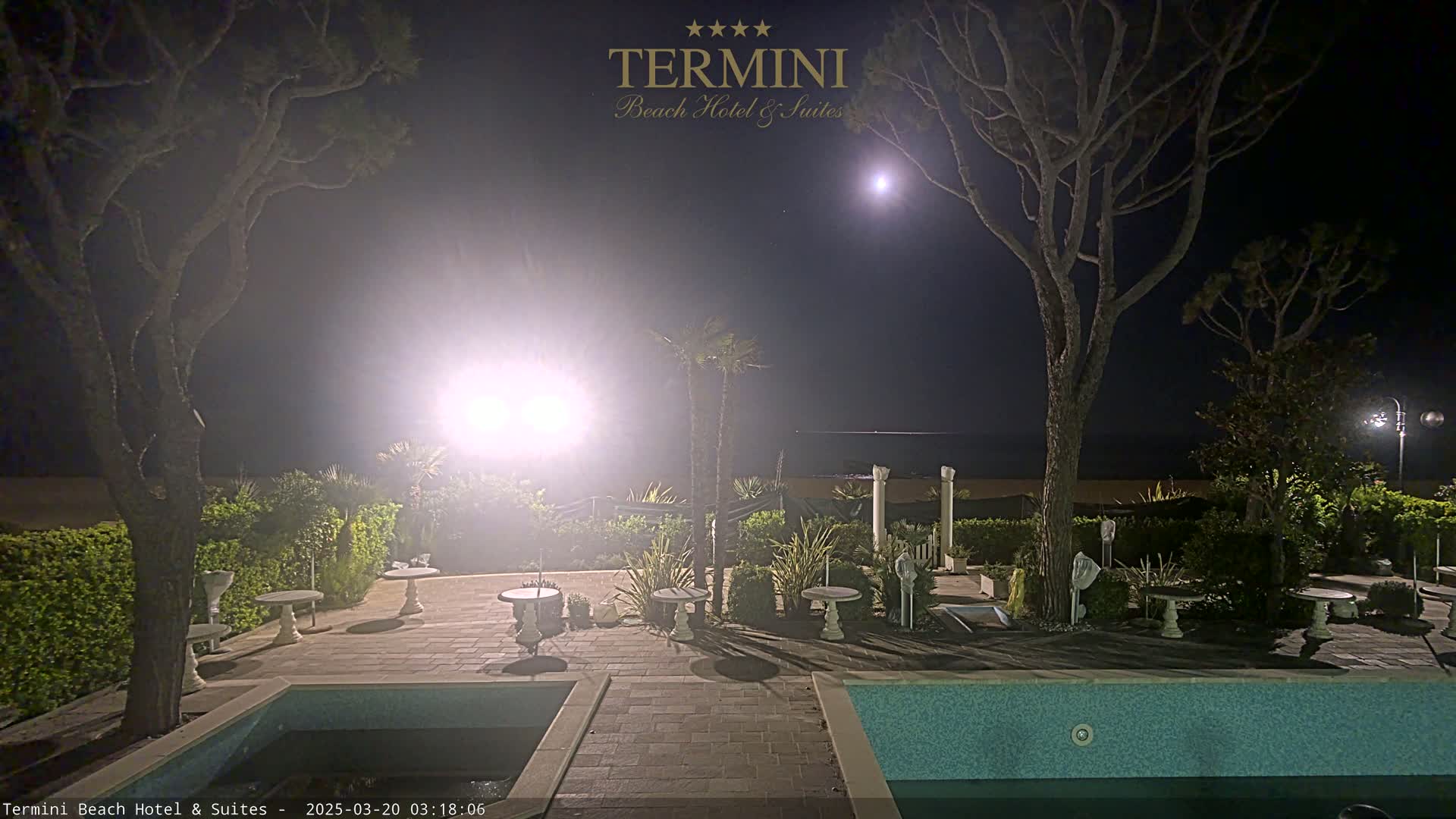 A nighttime view of a resort pool area, illuminated by lights, with trees and a moon visible in the clear night sky.