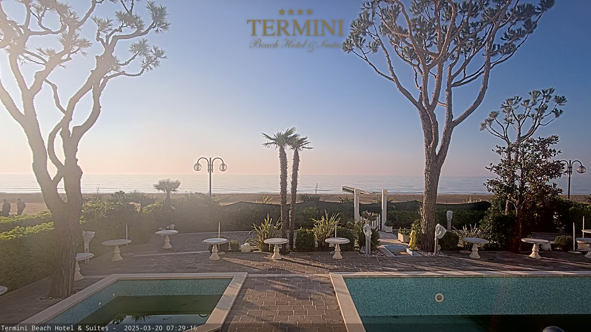 A courtyard with two swimming pools overlooks a calm ocean under a clear, bright sky.