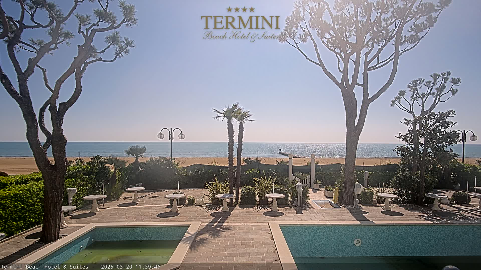 A sunny outdoor patio scene with two swimming pools, several small tables and chairs, palm trees, pine trees, and a view of a calm ocean and sandy beach.