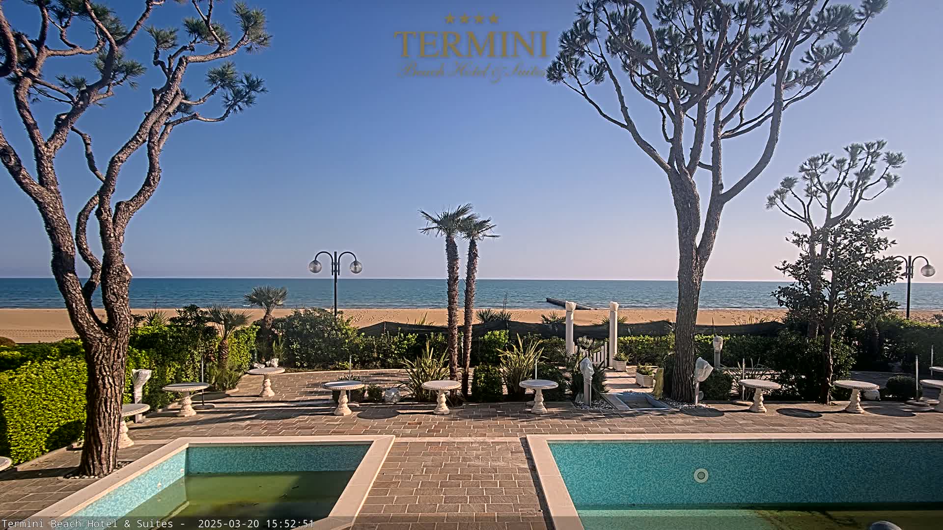 A tranquil outdoor patio scene features two pools, several small tables, and palm trees, all overlooking a calm ocean on a sunny day.
