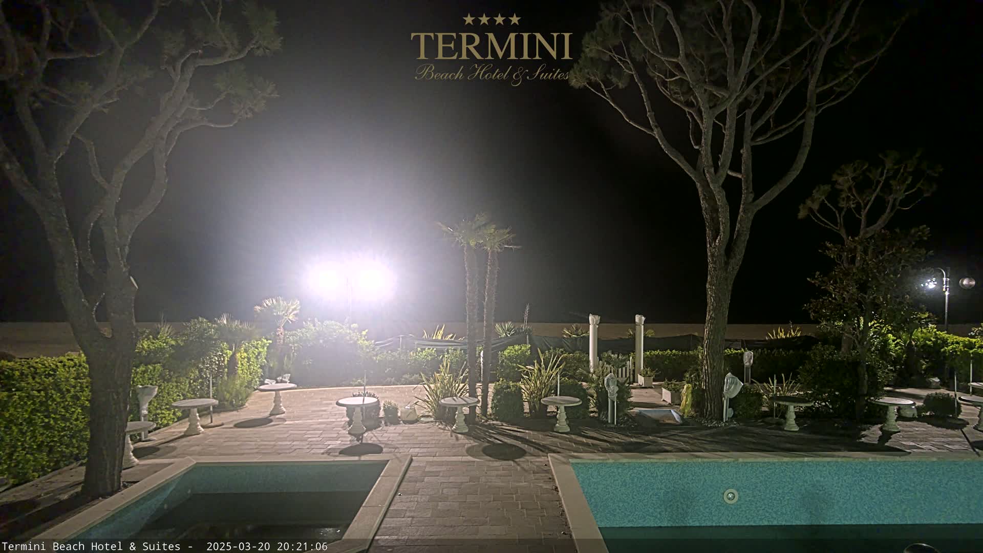 A nighttime view of a resort area with a swimming pool, patio furniture, and trees illuminated by bright lights.