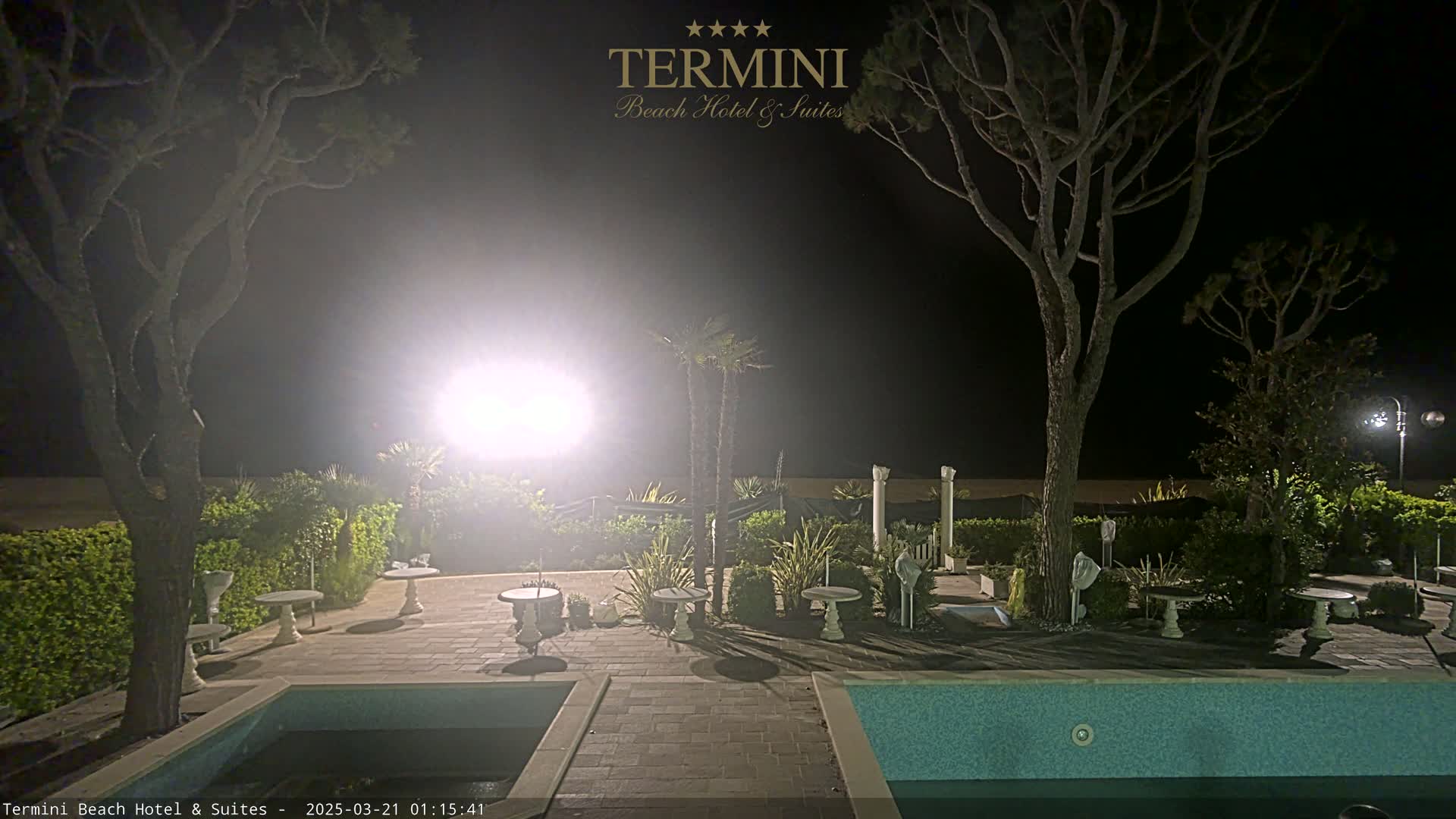 A nighttime view of a patio area with a swimming pool, surrounded by trees and landscaping, under bright outdoor lighting.