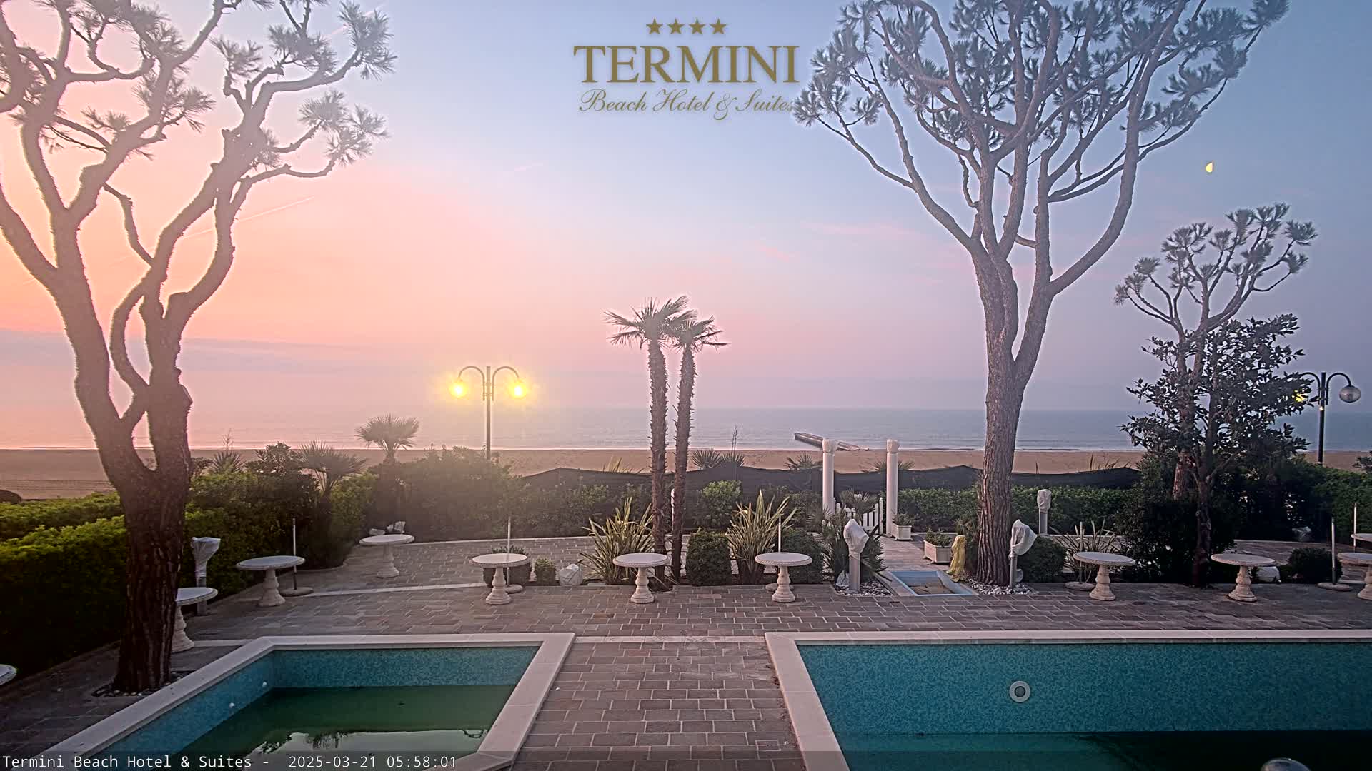 A tranquil outdoor scene at dawn shows a sandy beach and ocean under a pale pink sky, with two swimming pools and several small tables in a paved courtyard surrounded by trees and shrubbery.
