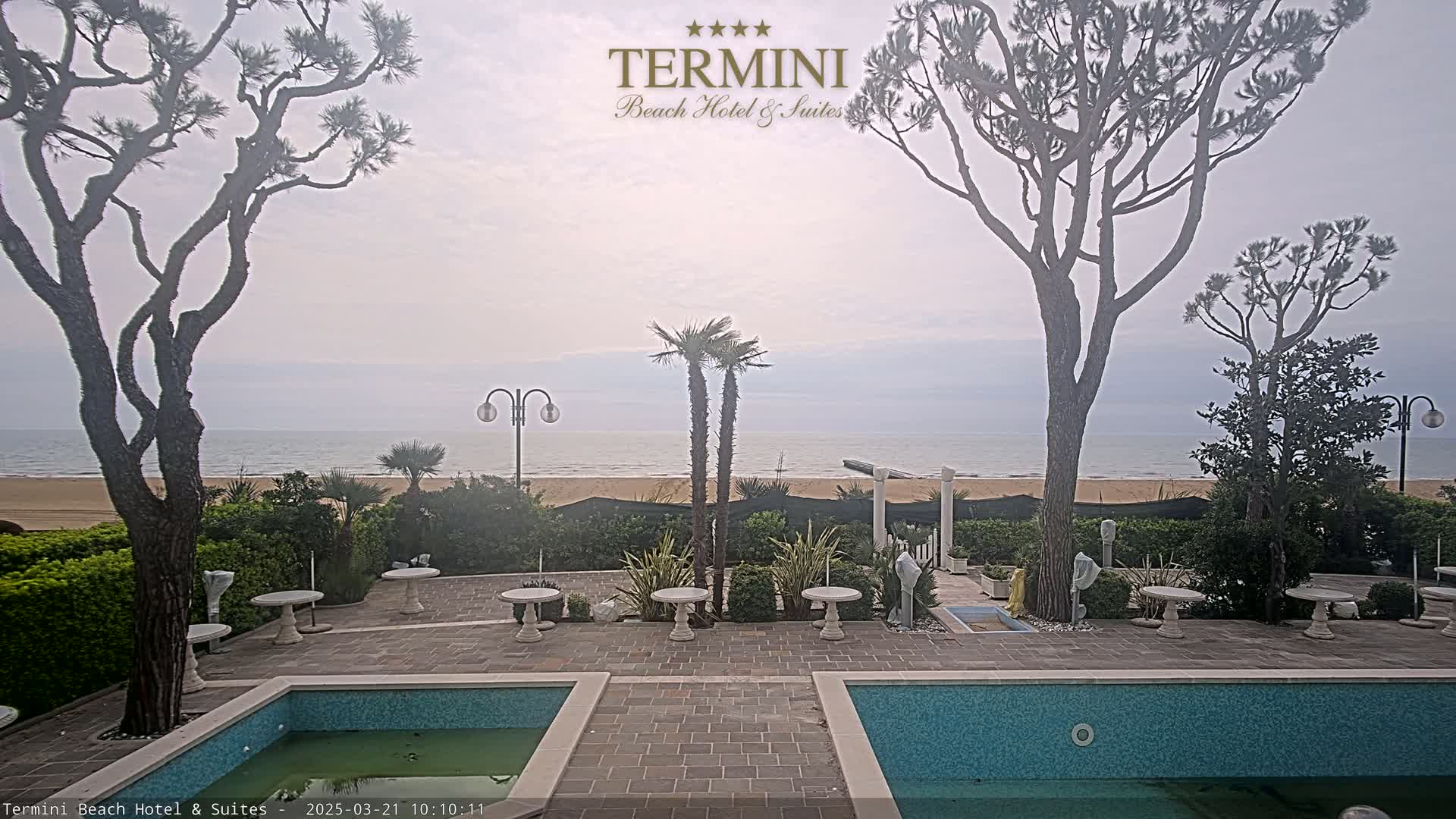 An overcast day shows a view of a sandy beach and calm ocean from a hotel courtyard with two swimming pools and landscaping.