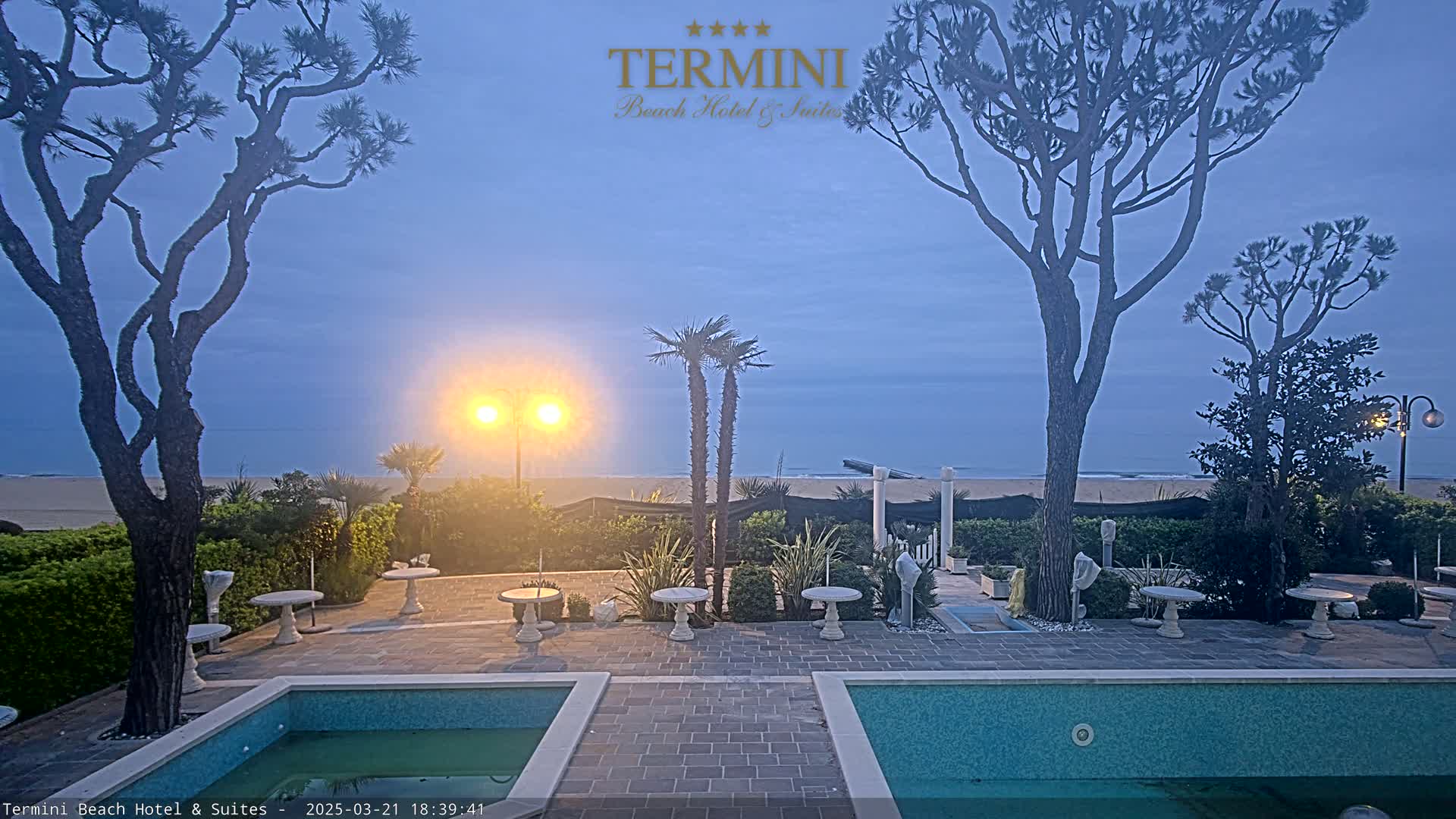 An outdoor patio area with two pools overlooks a calm, sandy beach at twilight under a clear, dusky sky.