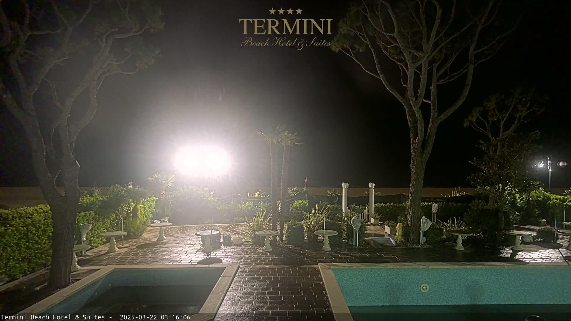 At night, a patio with two pools and several small tables is illuminated by bright lights under a hazy sky, surrounded by trees and shrubs.