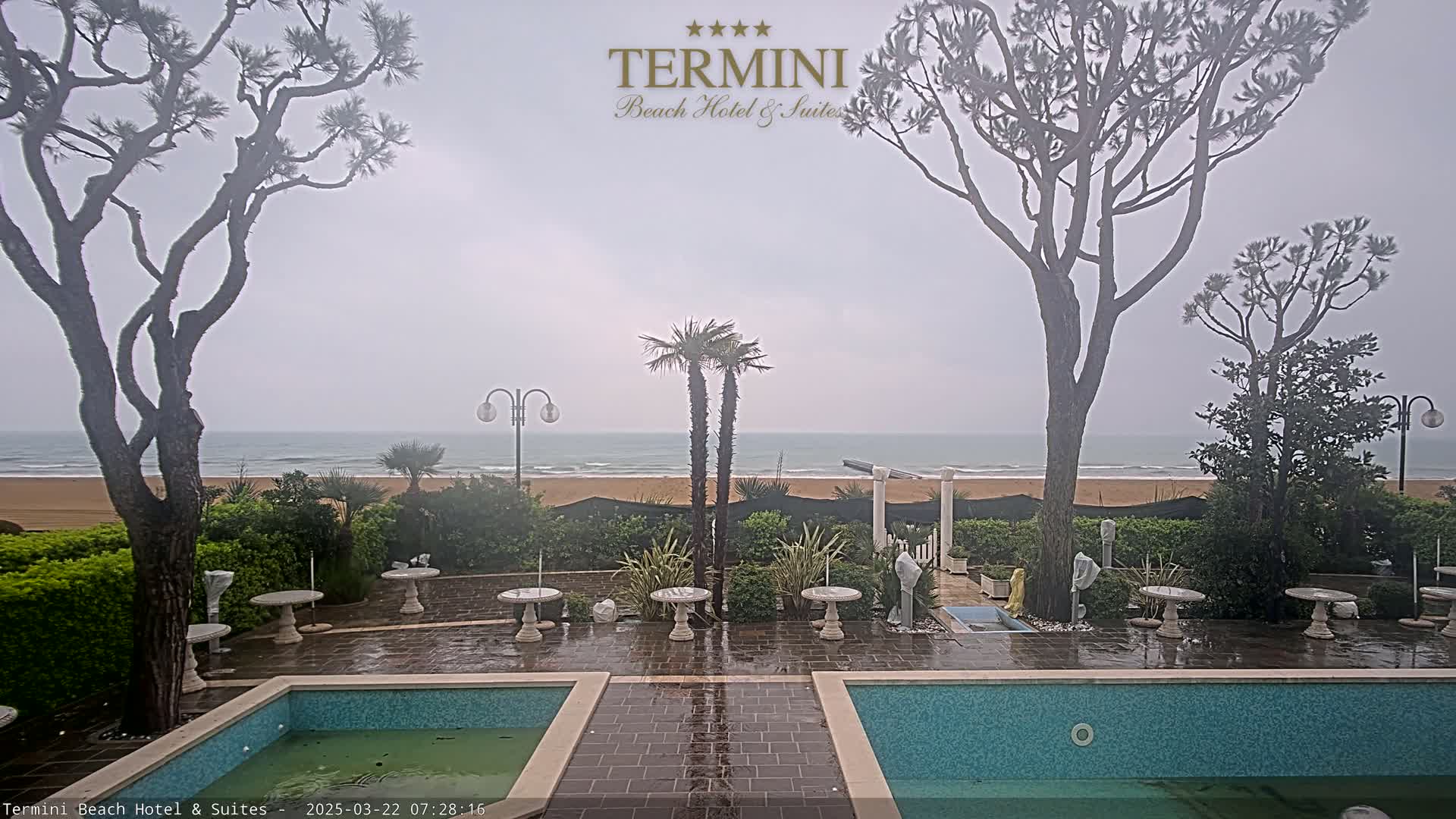 A patio with two pools overlooks a beach and ocean under an overcast sky.