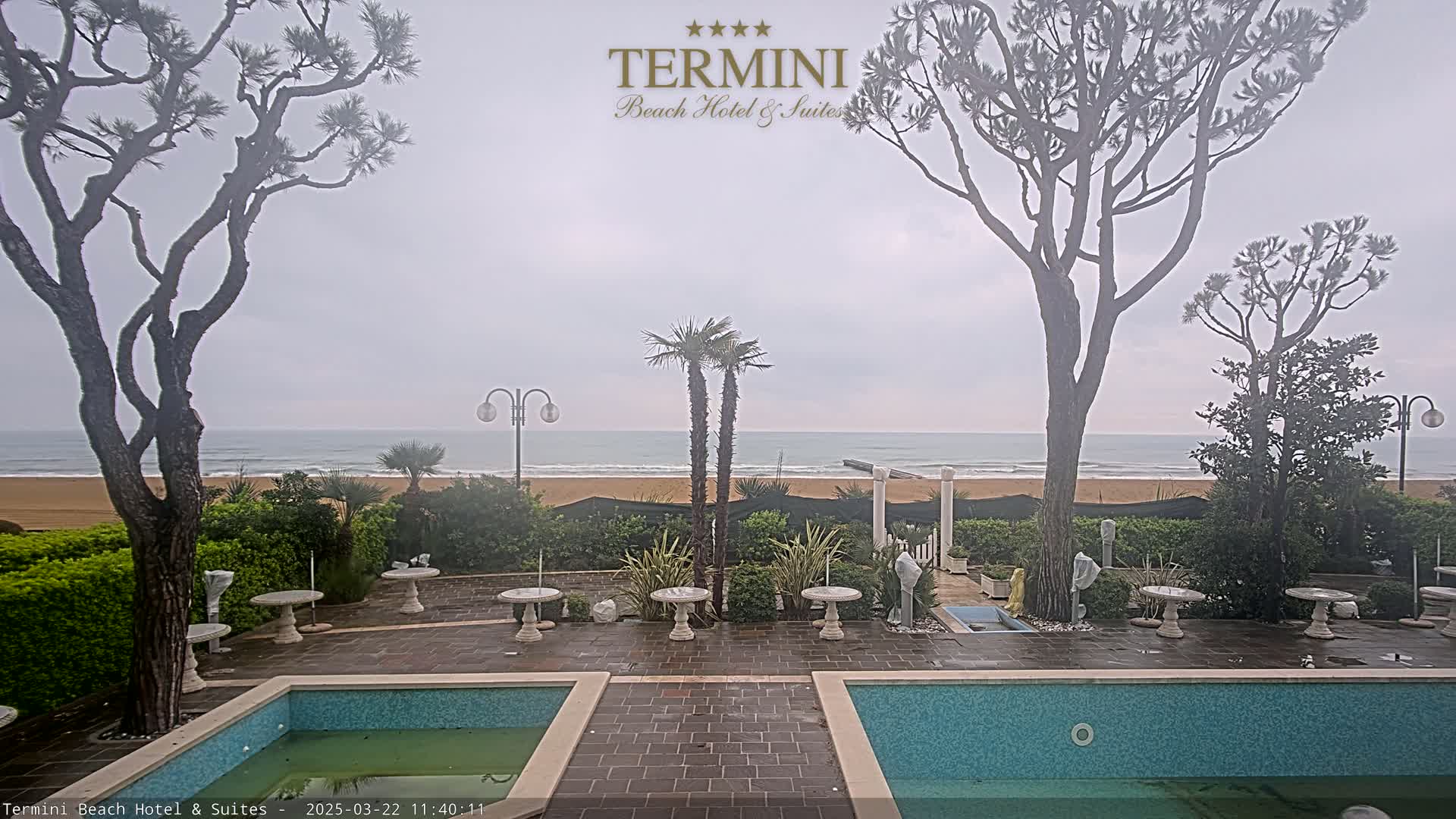 An overcast day shows a view of a sandy beach and ocean from a patio area with two small pools and several tables between trees and shrubs.
