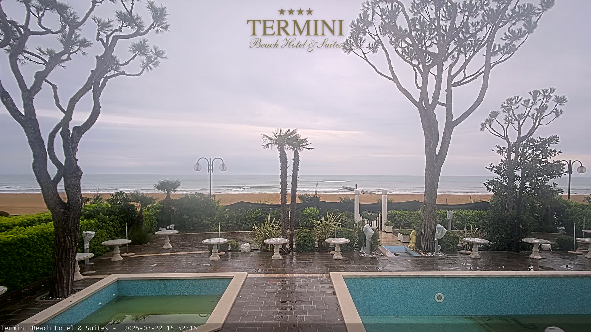 A patio with two pools overlooks a sandy beach and ocean under an overcast sky.