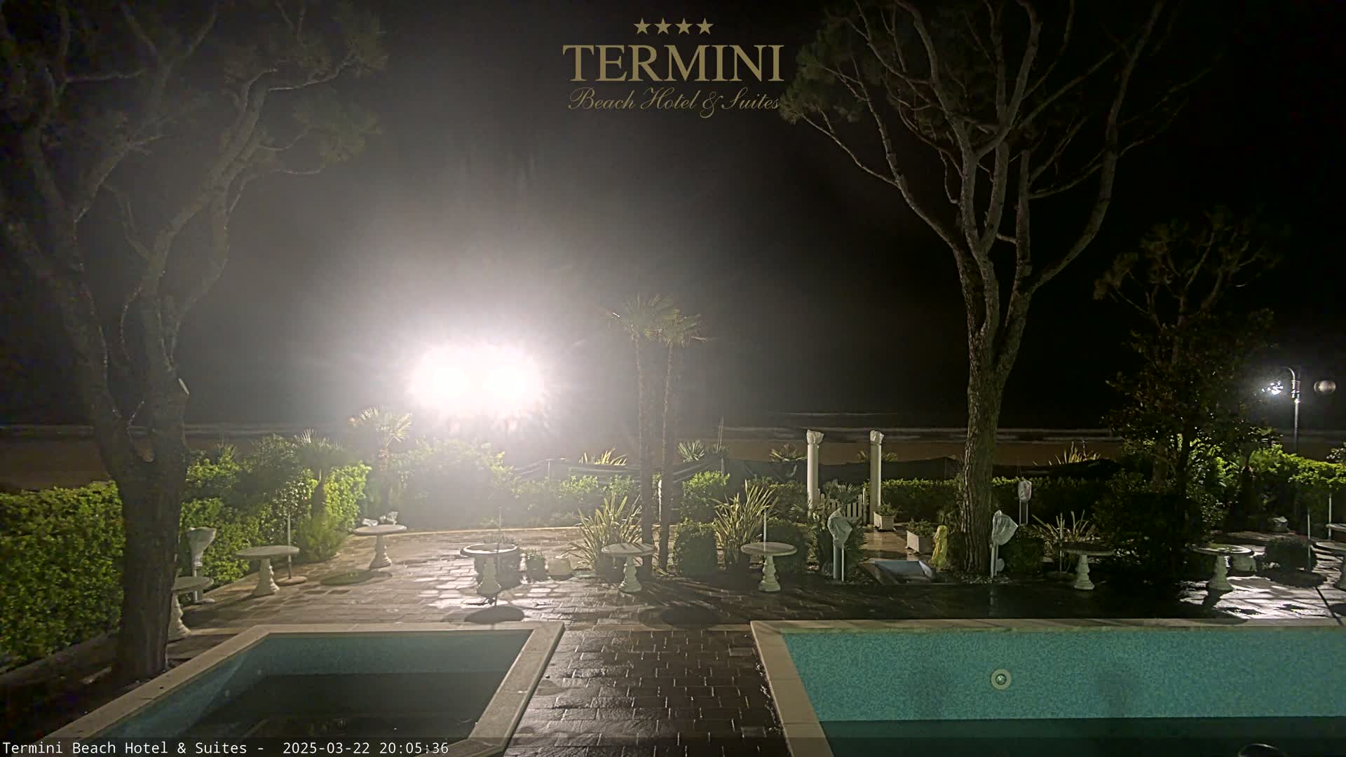 A nighttime view of a patio area with two swimming pools, several small tables, and trees, illuminated by lights in a hazy atmosphere.