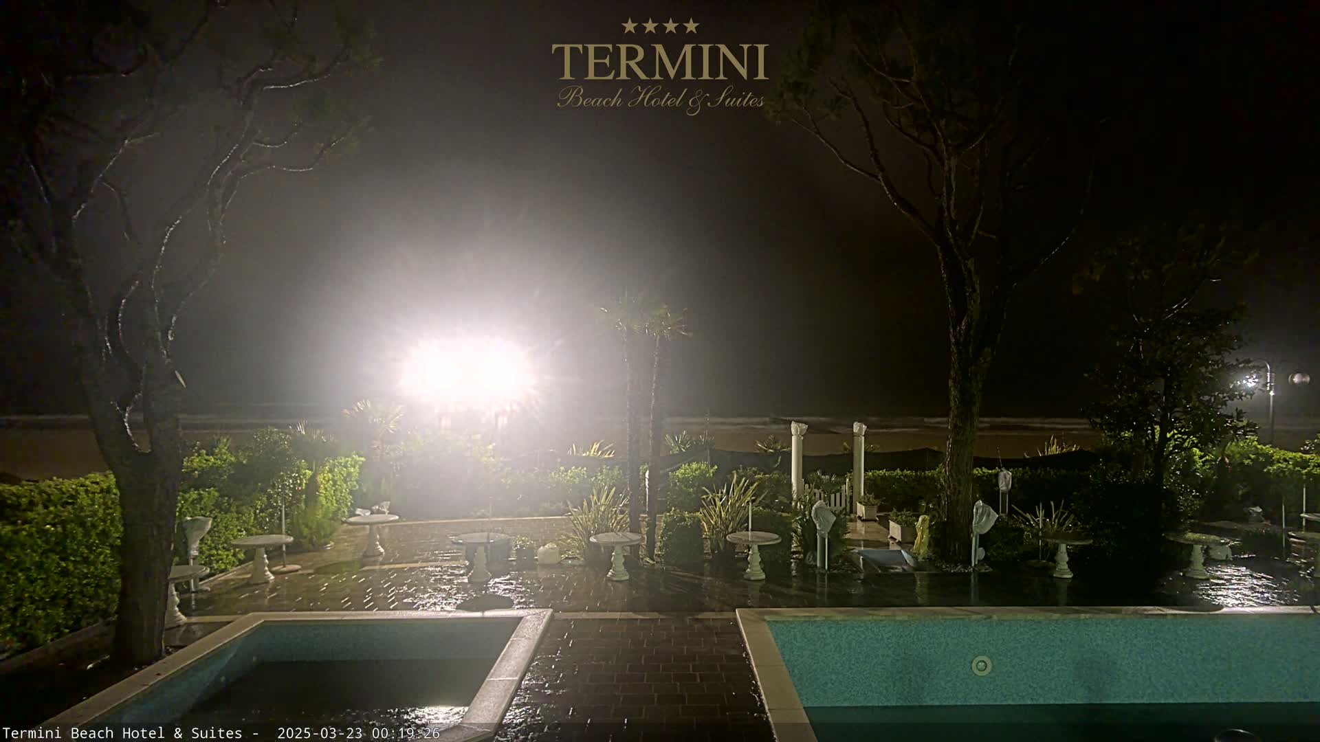 A nighttime view of a patio area with two pools, outdoor furniture, and landscaping, overlooking a foggy beach.