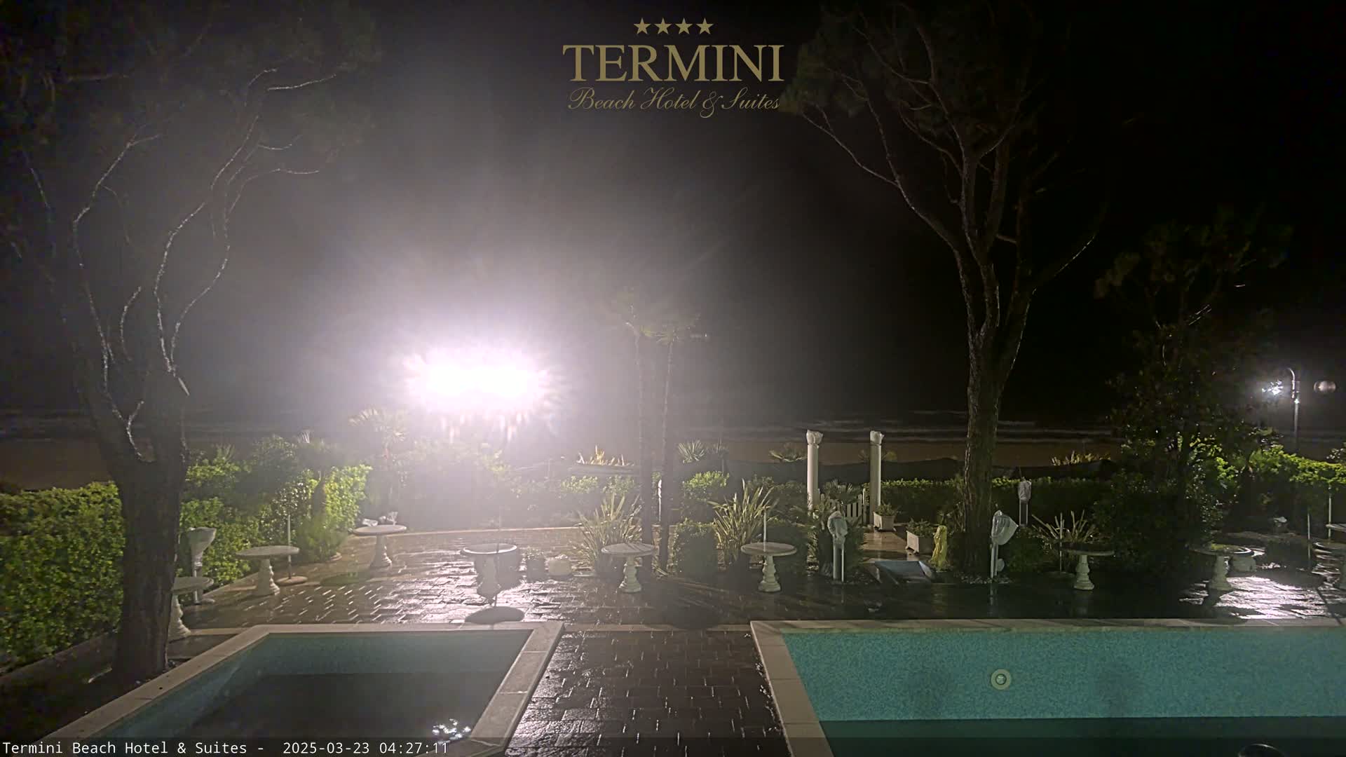 A nighttime view of a hotel patio with a pool, featuring wet patio furniture and trees under hazy, possibly foggy conditions.