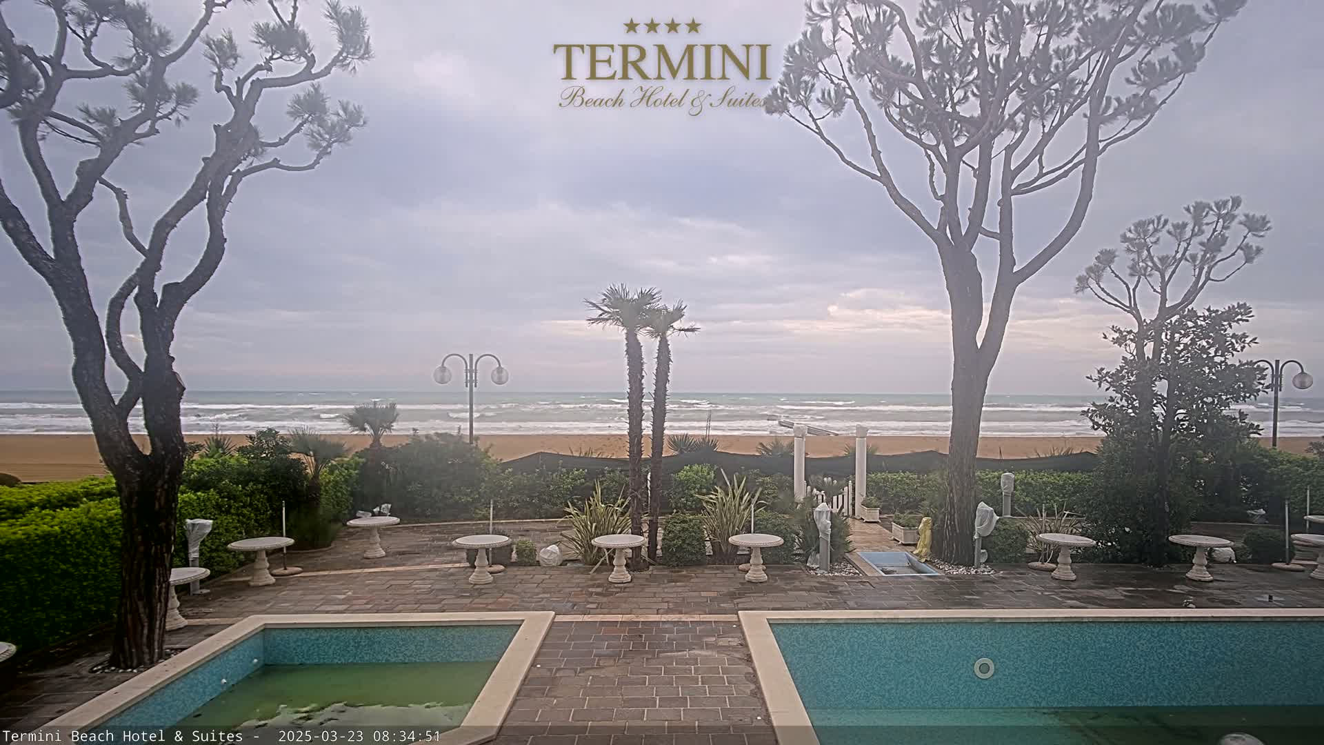 A patio area with two swimming pools overlooks a beach with wavy water under an overcast sky.