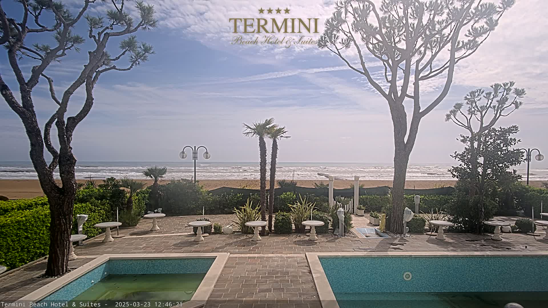 A patio with two swimming pools overlooks a sandy beach and a wavy ocean under a mostly sunny sky.
